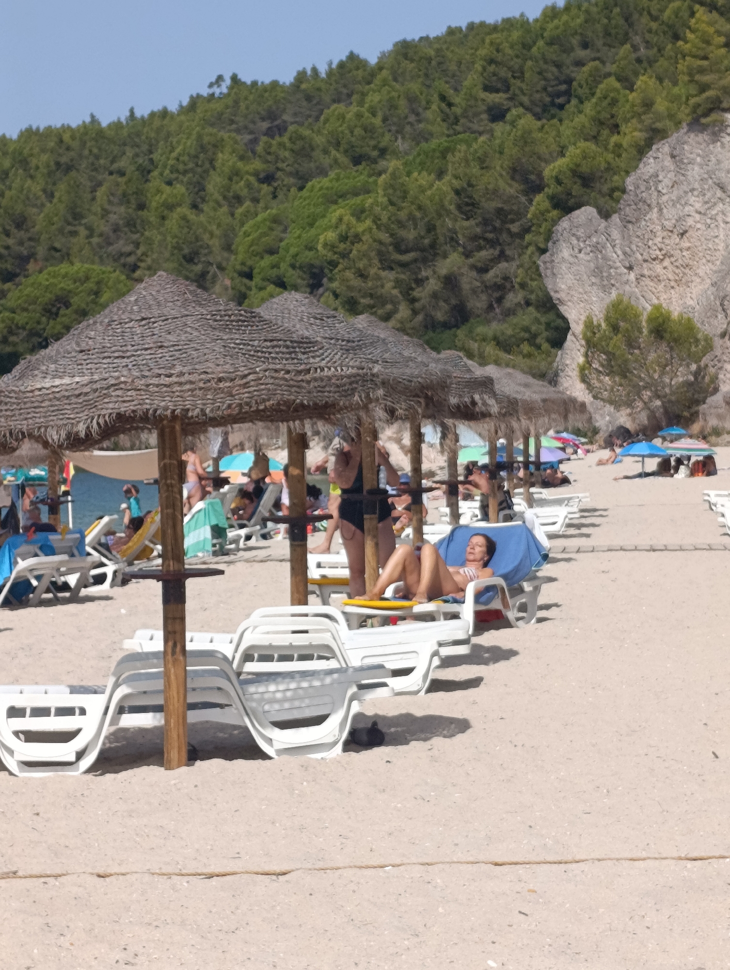 The image depicts a sunny day at a beach. In the foreground, several white plastic lounge chairs are stacked and arranged on the sand, some under thatched umbrellas. Further back, other beachgoers are visible, some lying on lounge chairs and others walking. In the background, a lush green hillside covered with trees rises behind the beach. The sea is visible in the distance. The overall scene conveys a relaxed atmosphere typical of a summer beach resort. The location is not specified as Setúbal, Portugal.