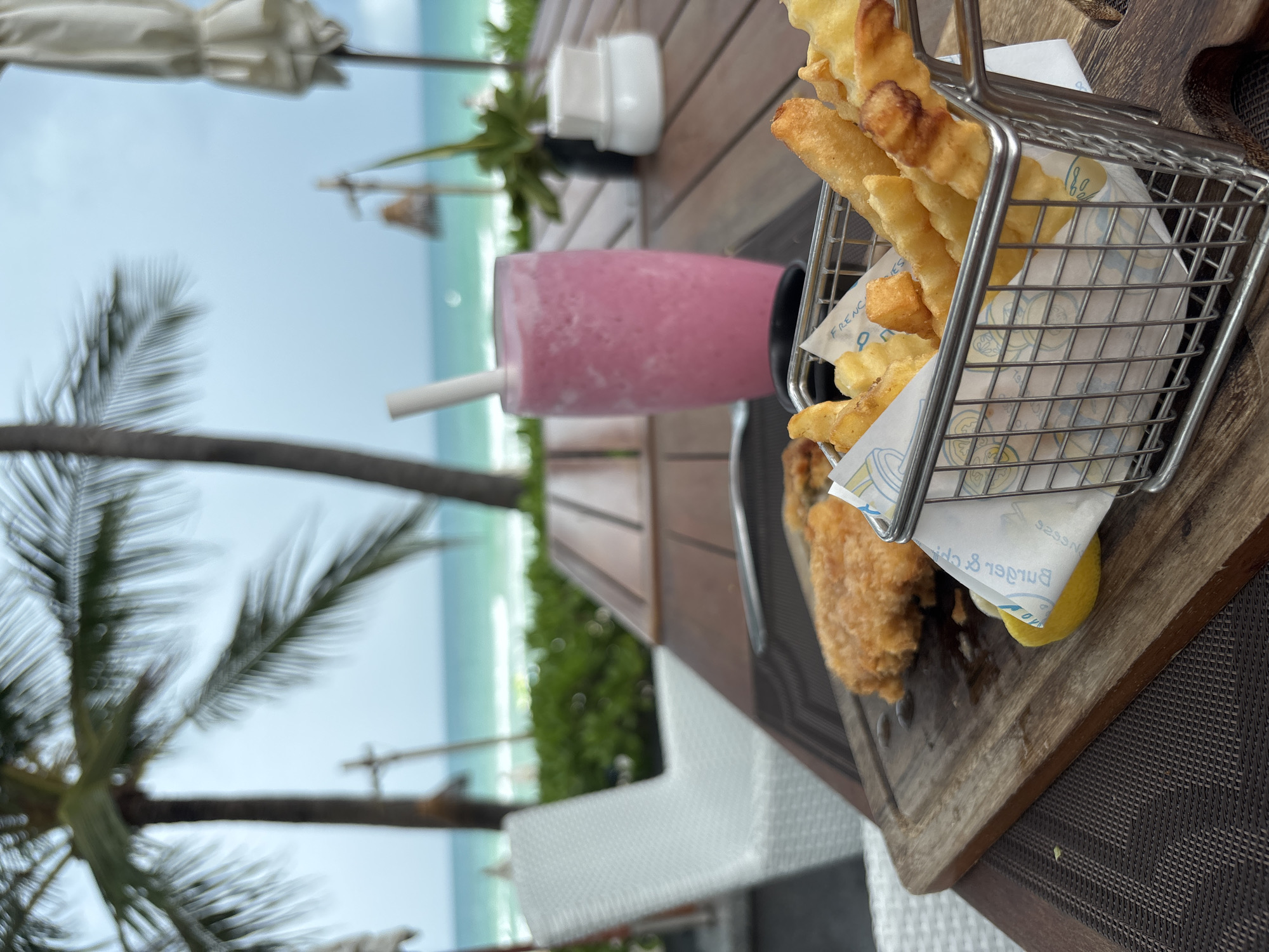 The image depicts a meal of fish and chips and a pink smoothie on a wooden table with a view of the ocean in Baan Chaweng, Thailand. The fish and chips are served on a wooden board with a wire basket for the fries. A lemon wedge is also present. A white straw is visible in the smoothie. A palm tree is visible, and the background contains a blue sea and sky. A white container is placed on the table as well.