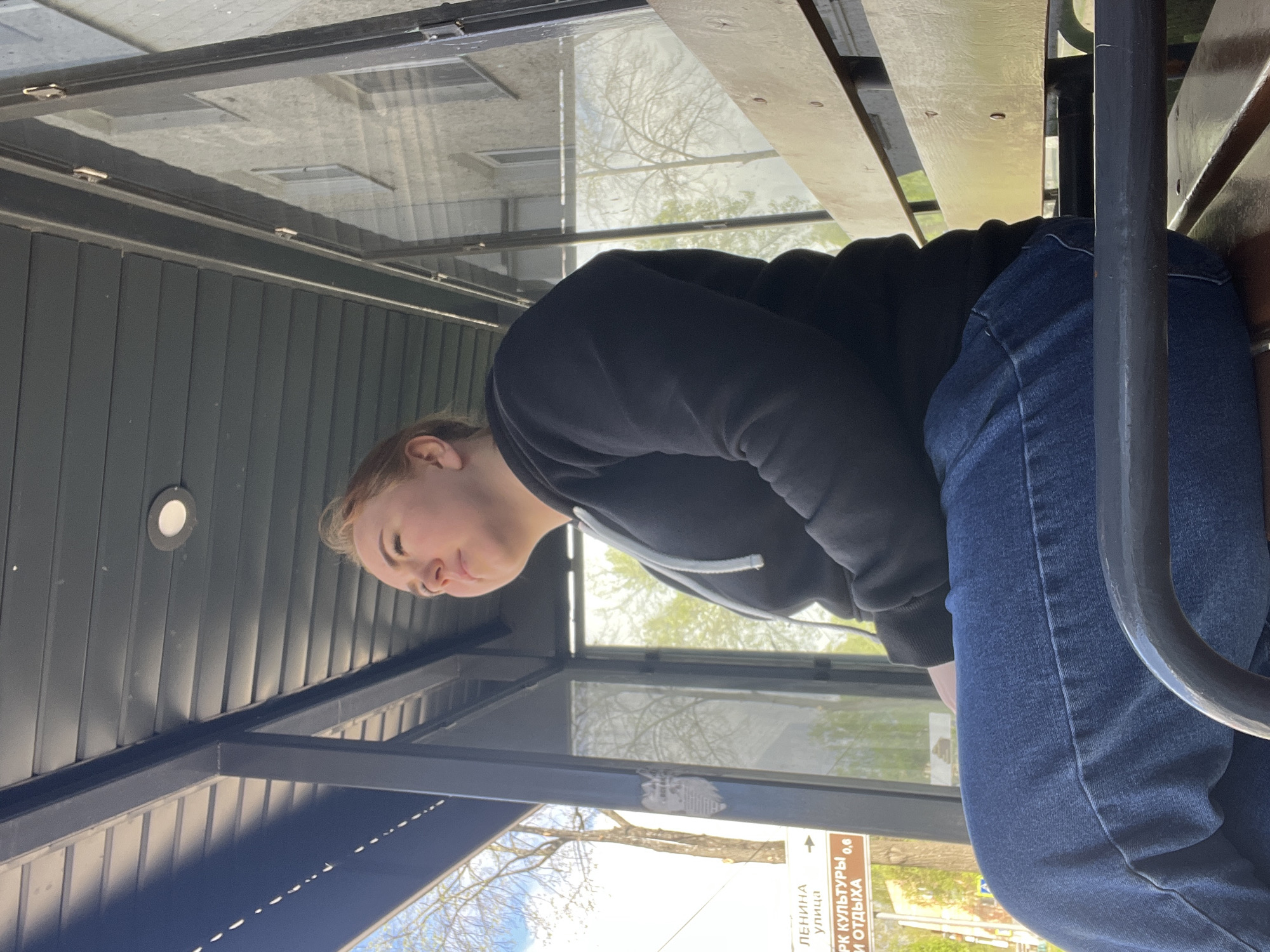 A person is seated on a wooden bench inside a bus stop in Chaikovsky, Russia. The person is wearing a black hooded sweatshirt and blue jeans. The bus stop has glass panels. Outside the bus stop, there is a sign in Russian. Trees are visible in the background.