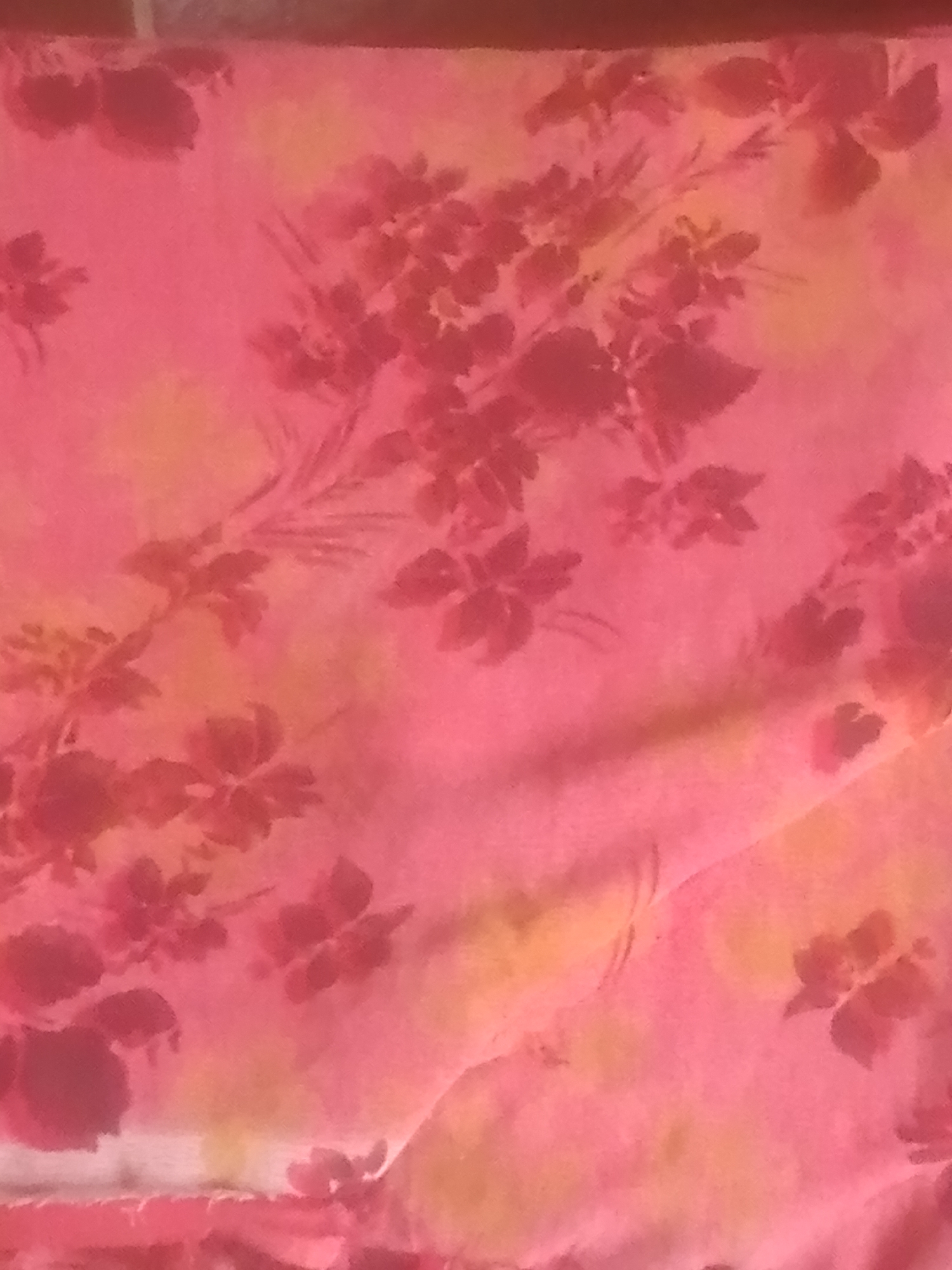 The image is a close-up, overhead shot of a pink fabric with a floral pattern. The flowers are depicted in a darker shade of pink and appear to be stylized or abstract. Some of the flowers have multiple petals, while others are more like clusters of leaves. There are also some yellow or pale orange splotches integrated into the fabric's design, creating a watercolor-like effect. The fabric appears to be soft and possibly silk or a similar material, with some subtle folds and creases visible, indicating it might be draped or spread out. The lighting is soft and diffuse, giving the image a gentle and somewhat dreamy quality. The specific location within Jauro Yinu, Nigeria, cannot be determined from this image alone, as it focuses solely on the texture and pattern of the fabric.