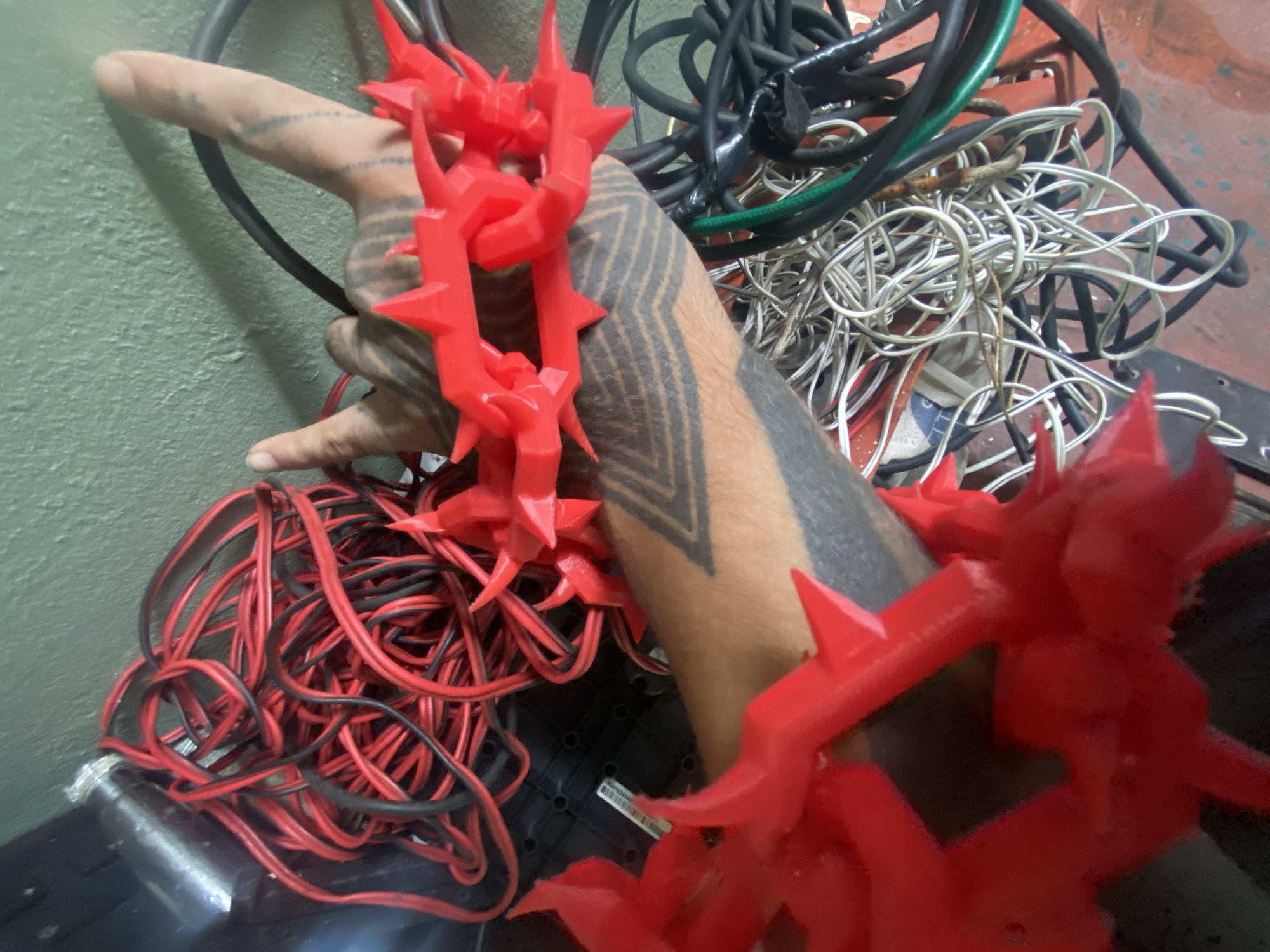 An adult arm with tribal tattoos is draped with a red, spiked, plastic chain. The arm is surrounded by tangled wires of various colors, including red, black, white, and green. The scene is cluttered and appears to be indoors, potentially a workshop or storage area. The location in Buenos Aires, Argentina, is not discernible from the image.