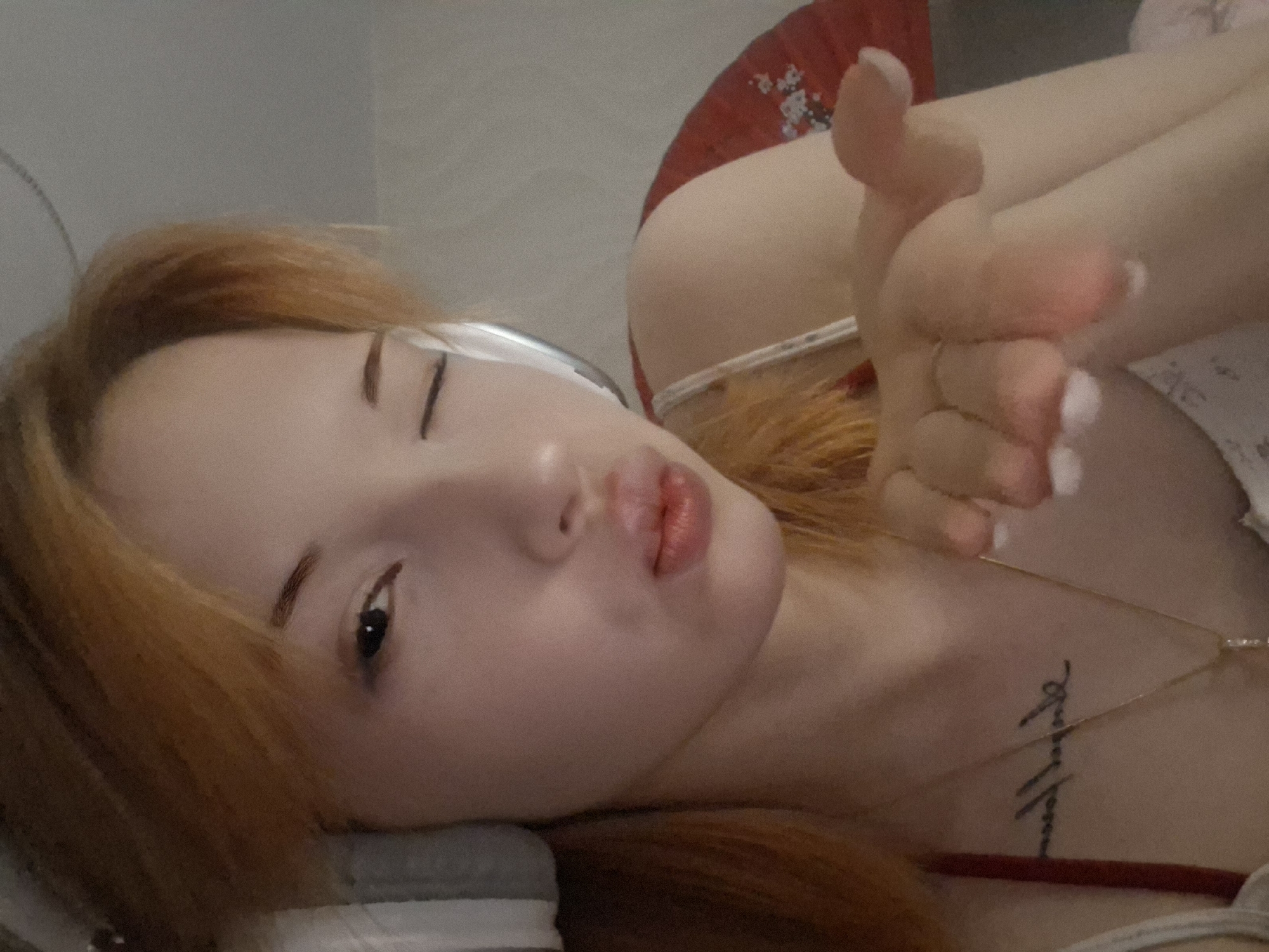 A young woman with auburn hair is lying down, winking one eye, and puckering her lips. She is extending her hand with fingers curled inwards. A necklace is visible around her neck, and a tattoo is visible on her chest. A red and white decorative fan is positioned in the background, partially visible behind her shoulder. The environment is a bedroom. There is no information that allows for the specification of the exact location in the city of Chaikovsky, Russia.