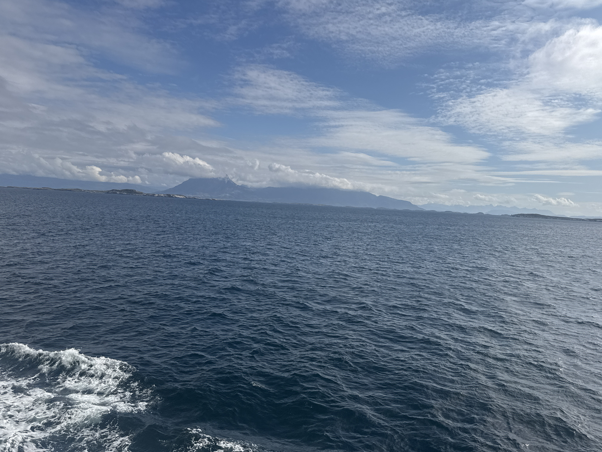 The image depicts a vast expanse of dark blue ocean under a partly cloudy sky. The water is choppy, with white foam and waves visible in the foreground on the left side of the frame, indicating movement. In the distance, a mountainous coastline stretches across the horizon, with some peaks obscured by clouds. The sky is a mix of bright blue patches and white, wispy clouds, with some larger, more defined clouds casting shadows on the distant mountains. The overall impression is one of a rugged, natural landscape. The specific location in Nordsolvær, Norway is a coastal area characterized by its maritime environment, mountainous terrain, and open sea. There are no people or man-made objects visible in the image.