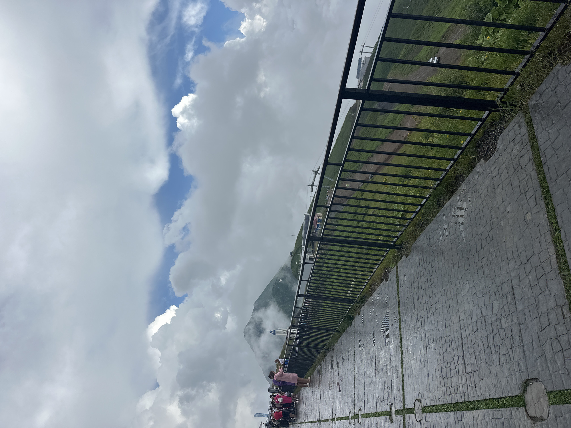 An outdoor, eye-level, full shot captures a paved walkway with a black metal railing overlooking a steep, grassy mountain slope under a cloudy sky. The walkway is constructed from large, dark grey, rectangular pavers with grass growing in the gaps between them, and appears wet, with visible puddles reflecting the sky. A group of people, some dressed in casual clothing including a woman in a pink dress, are walking and standing on the walkway on the left side of the image. The mountain slope to the right is covered in lush green grass with patches of yellow wildflowers. Further down the slope, a dirt road is visible with a white car driving on it, and several buildings and ski lift structures are scattered on the mountainside. In the background, a mist-shrouded mountain peak rises towards the sky, partially obscured by dramatic white and grey clouds. The scene is characteristic of a viewpoint at a mountain resort in Estosadok, Russia, likely during the summer or shoulder season given the green vegetation and cloudy weather.