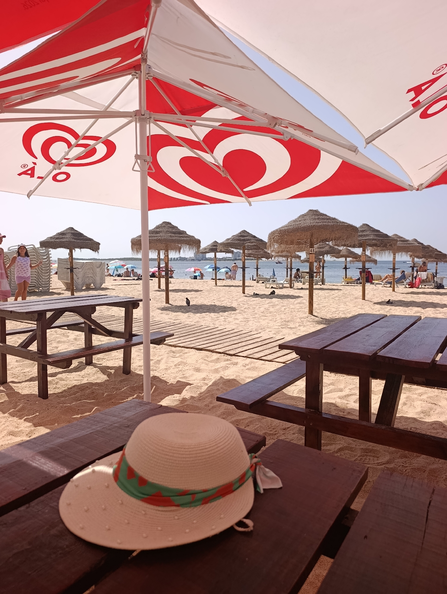 The image depicts a sunny day on a sandy beach in Setúbal, Portugal. In the foreground, a picnic table with a straw hat on it is visible, under a large red and white striped umbrella. The umbrella appears to be branded with a red swirl logo. In the middle ground, several thatched umbrellas are spread across the sand, with beach chairs and umbrellas in the background. People are scattered across the beach, some sitting, some standing, and some wading in the calm water. The sea is visible in the distance, with a few boats on the horizon. The overall scene suggests a relaxed vacation atmosphere.