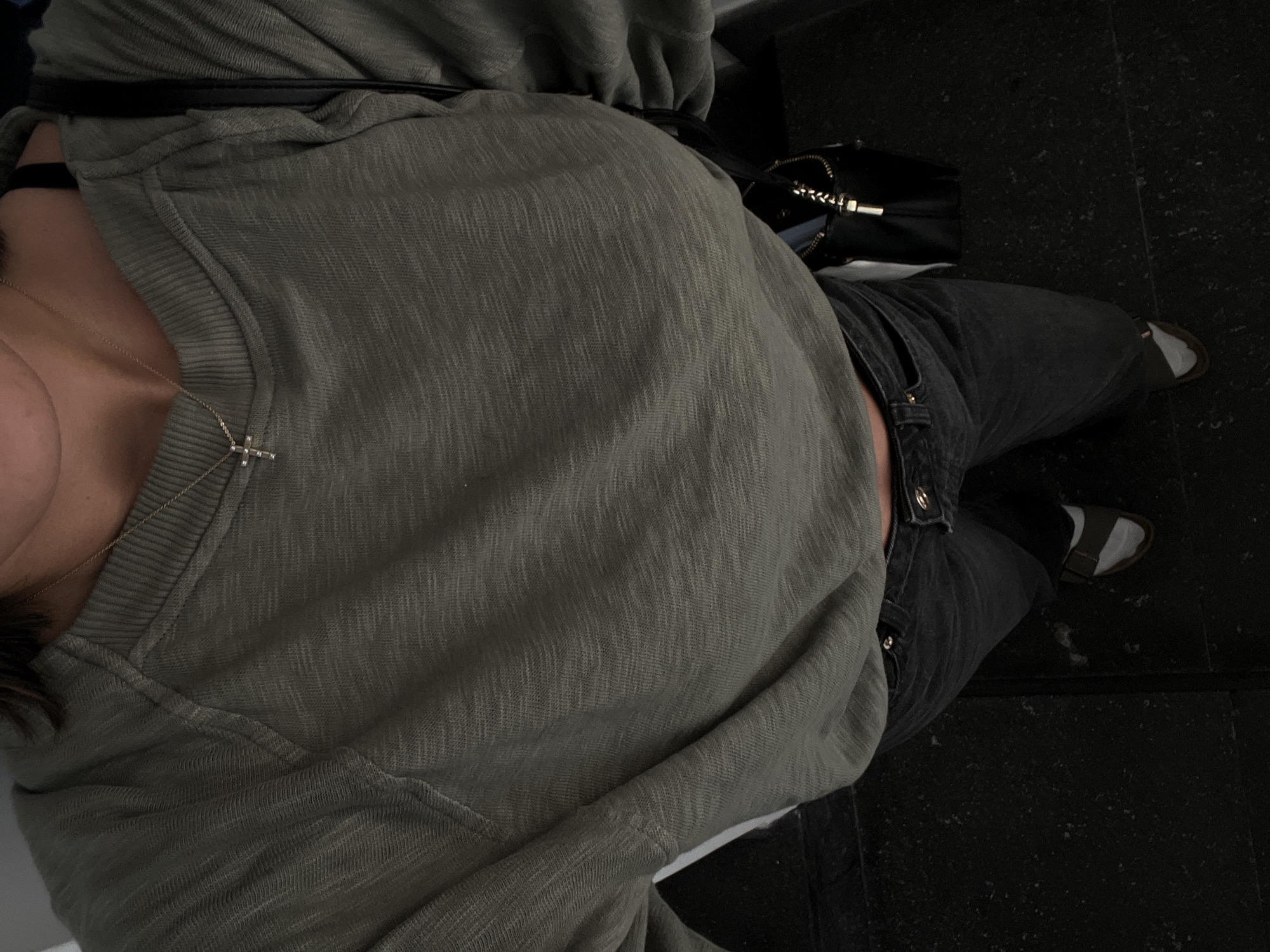 A high-angle medium shot shows a person wearing a green long-sleeved shirt, black jeans, white socks, and sandals, standing on a dark textured floor that appears to be indoors, possibly a staircase. A black purse with a gold zipper hangs from the person's right shoulder. The person is also wearing a delicate silver necklace with a cross pendant. The lighting is dim, with shadows obscuring much of the background and floor details. The scene is located in San Bernardino Tlaxcalancingo, Mexico.