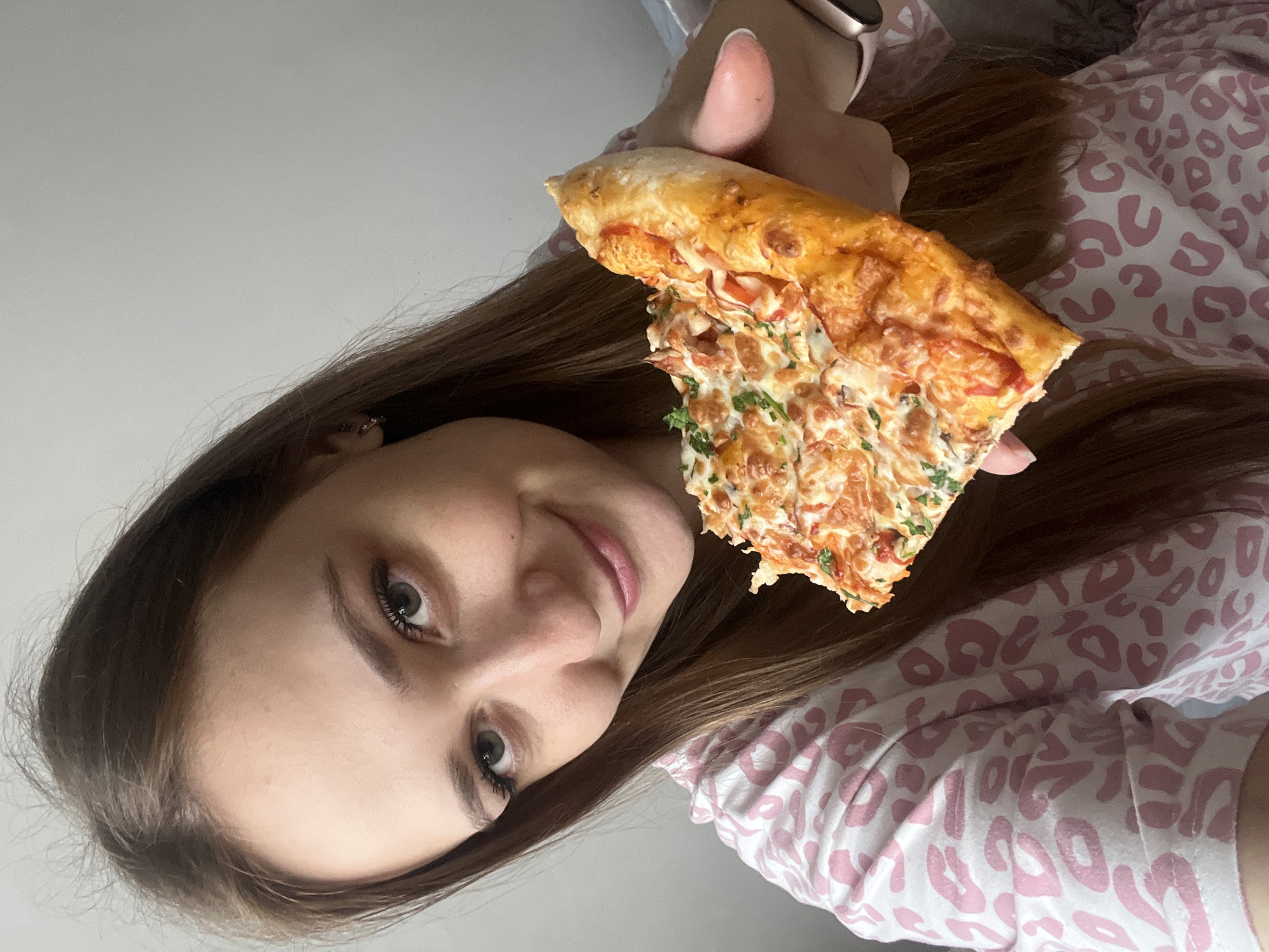 A young woman with long brown hair holds a slice of pizza in her right hand. The pizza appears to be a combination of two different toppings: one side has a red sauce with what appears to be sausage and cheese, and the other side has a creamy white sauce with diced tomatoes, parsley, and possibly chicken or other small pieces. She is wearing a long-sleeved shirt with a pink leopard print pattern. The image appears to have been taken indoors. The location is not apparent from the image and there is nothing to suggest that it was taken in Verkhnyaya Pyshma, Russia.