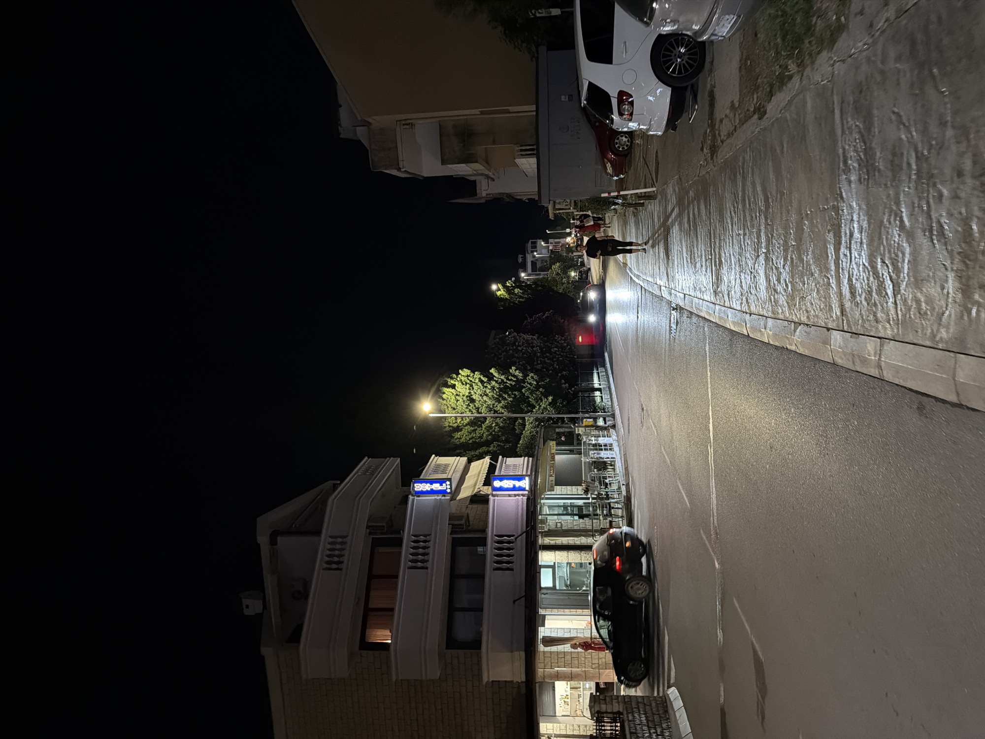 The image depicts a nighttime street scene in Budva, Montenegro. The street is paved and illuminated by streetlights and lights from buildings. On the left side of the street, a multi-story building with balconies and a sign that reads "HOTEL" is visible. Below the hotel, there is a business with large glass windows, and a black car is parked in front of it. A person in a red top is standing inside this establishment. Further down the street, trees are illuminated by streetlights. On the right side of the street, a sidewalk is present, with several cars parked along it, including a white SUV and a red car. A woman in dark clothing is walking on the sidewalk, facing away from the camera. In the background, other buildings and trees are visible, as well as a few other pedestrians and vehicles. The overall atmosphere is that of a quiet evening in a coastal town.