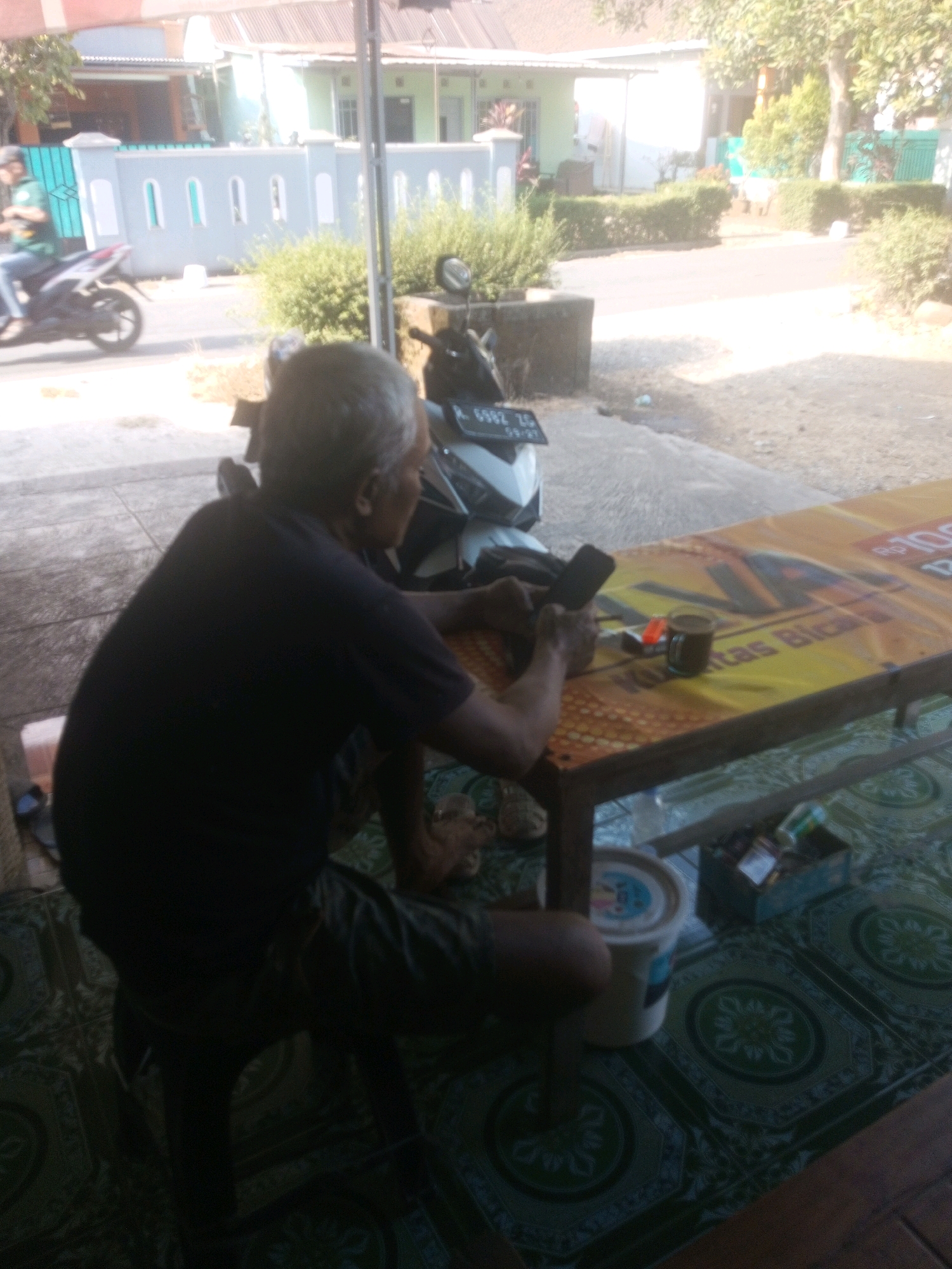 A low-angle, medium shot captures an elderly man with graying hair sitting on a stool and looking down at a smartphone in his right hand. He is wearing a dark t-shirt and dark shorts. His left hand rests on a table covered with a colorful tablecloth featuring Indonesian text and images. On the table, a glass of dark liquid, a pack of cigarettes, and a lighter are visible. The man is positioned in front of a building with a light-colored roof and walls, and a turquoise-colored fence with arched openings. A motorcycle is parked behind him. The scene appears to be outdoors, with trees and vegetation in the background. The floor is tiled with a repeating green pattern. The overall lighting suggests it is daytime, with harsh sunlight creating some overexposed areas. The image quality is slightly blurred.