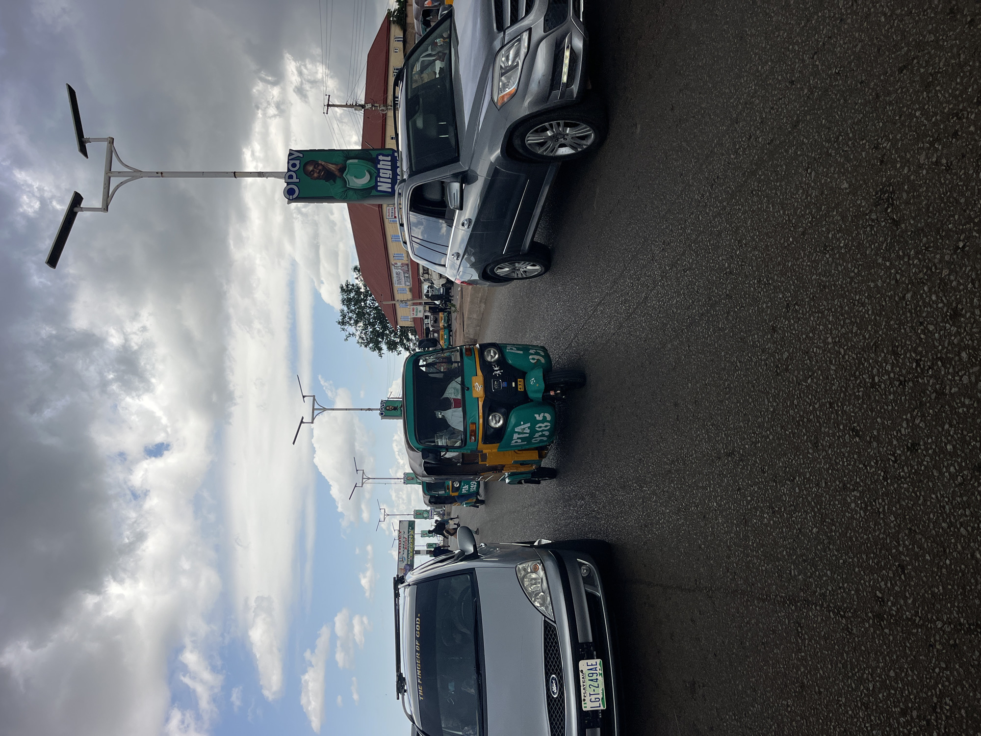 A low-angle, medium shot captures a street scene in an unknown city, likely in Africa, under a cloudy sky. The foreground features the front of a silver Ford Focus, its license plate reading "LGT-249AE" with "PLATEAU" above it, and a sticker on the windshield saying "THE FINGER OF GOD." To its right, a grey Mercedes-Benz GLK-Class SUV is visible. Dominating the middle ground are several three-wheeled auto-rickshaws, or "keke napep," painted in bright green and yellow, with one clearly marked "PTA 9385." People are visible inside and around the vehicles, suggesting active traffic. In the background, buildings with red roofs line the street, and several modern streetlights equipped with solar panels are prominently displayed. One streetlight has a green advertisement banner featuring a man and the word "OPay" and "Night." The road surface is asphalt, appearing wet in places. The overall impression is of a bustling urban environment with a mix of vehicles and infrastructure.