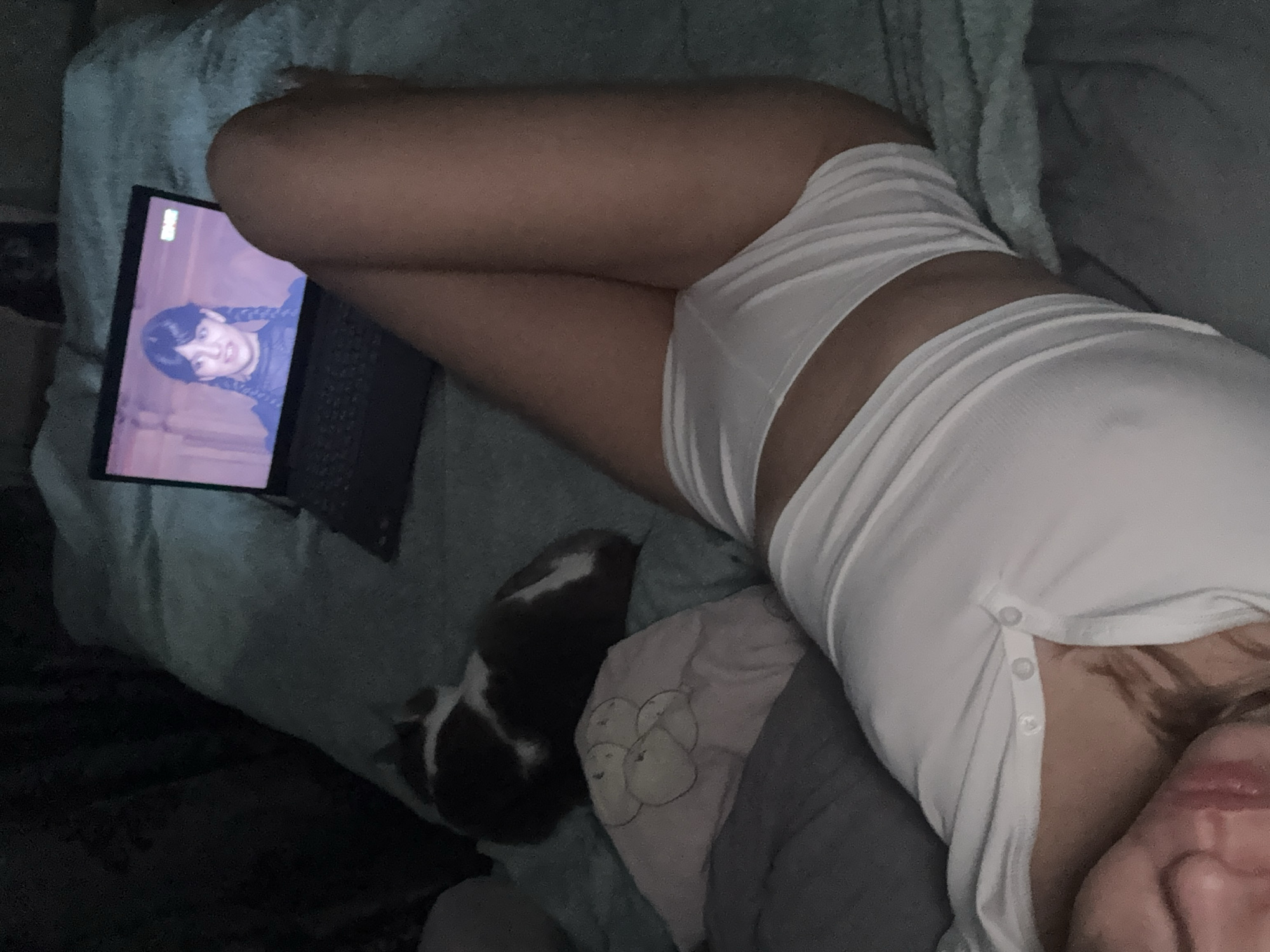 The image captures an individual relaxing indoors, lying on their side on a bed or couch in dim lighting, likely during the evening or night. The person is wearing white, ribbed loungewear, consisting of shorts and a top with visible buttons along the side, and their legs are bent in a relaxed posture.
A laptop is positioned beside their legs, displaying a character on the screen with dark braided hair, strongly resembling Wednesday Addams, suggesting a television show or movie is being watched. Faint, illegible text is visible in the top left corner of the laptop screen, likely a logo from a streaming service.
A small pet, possibly a guinea pig or a rabbit, with patches of dark and white fur, is nestled on the light blue/grey textured bedding between the person and the laptop. Another pillow with a subtle, light-colored fruit design is also partially visible.
The overall scene conveys a calm and casual home environment. The low light contributes to a cozy atmosphere. The person's face is largely out of frame, so emotions are not discernible. No specific visual cues within the image indicate the geographical location as Ust-Ilimsk, Russia.
