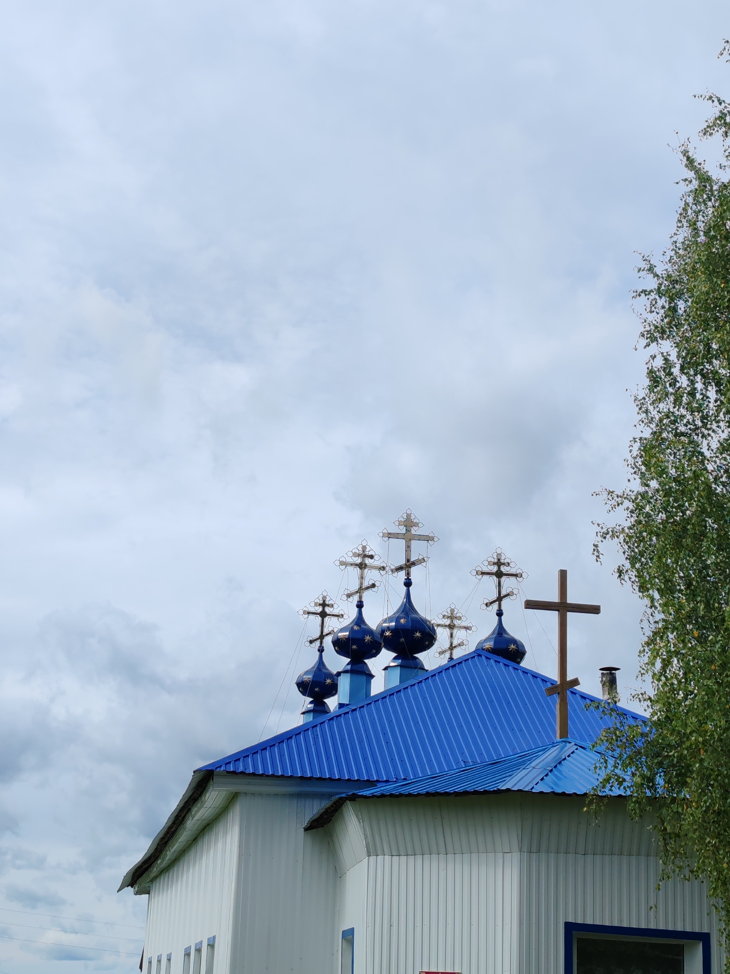 The image shows the top section of a white, tin-clad building with a bright blue, corrugated metal roof. Several blue onion domes with gold star accents are visible on the roof, each topped with an ornate cross. A large, simple wooden cross stands prominently to the right of the domes. The background consists of a cloudy, pale blue sky. To the right of the building, the green leaves and branches of a tree are visible, partially obscuring the sky. The building appears to be a church or a religious structure. The scene is set outdoors, under an overcast sky.