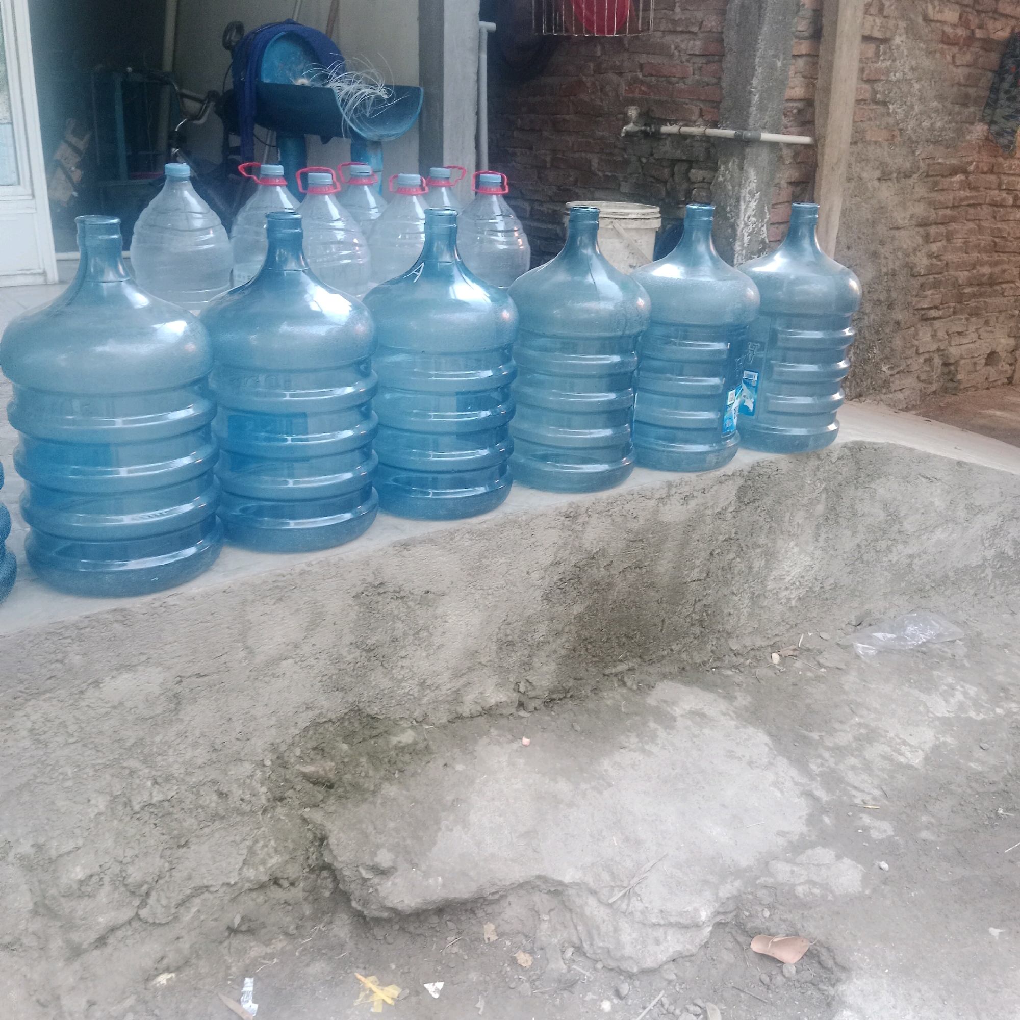 In Tegowanu Kulon, Indonesia, a row of large, blue plastic water jugs are lined up on a concrete surface in front of a brick building. The jugs are filled with water and have white or red caps. Some jugs have labels visible. The background shows a brick wall, a pipe with a faucet, and some debris on the ground. The scene appears to be an outdoor storage area or a water refill station.