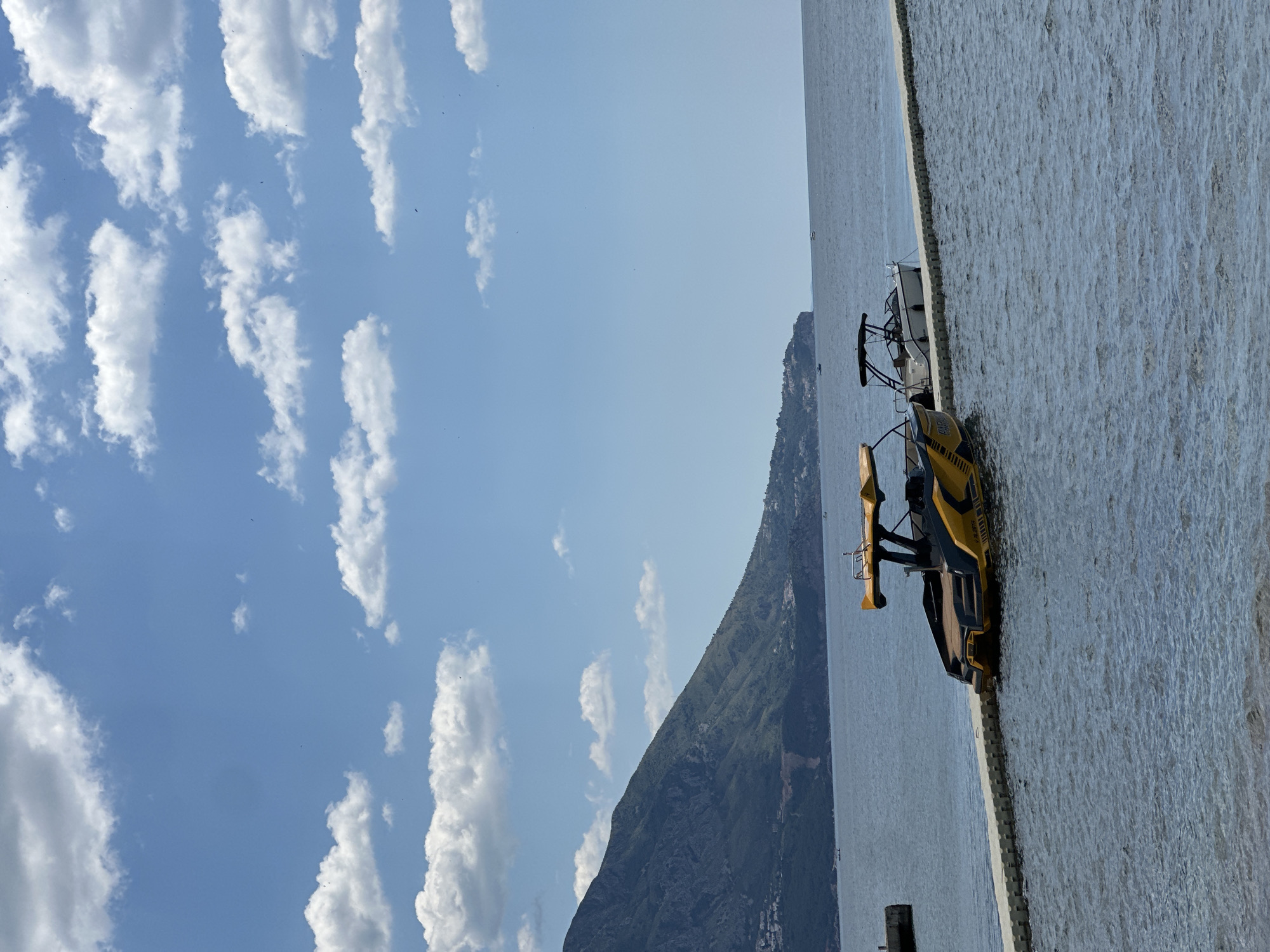 The image captures a scenic view in Budva, Montenegro. The foreground features a yellow and black boat docked on a waterfront pier. The backdrop showcases a mountainous landscape meeting the Adriatic Sea under a bright blue sky dotted with scattered white clouds. The sea appears calm and stretches to the horizon. The image conveys a sense of tranquility and natural beauty.