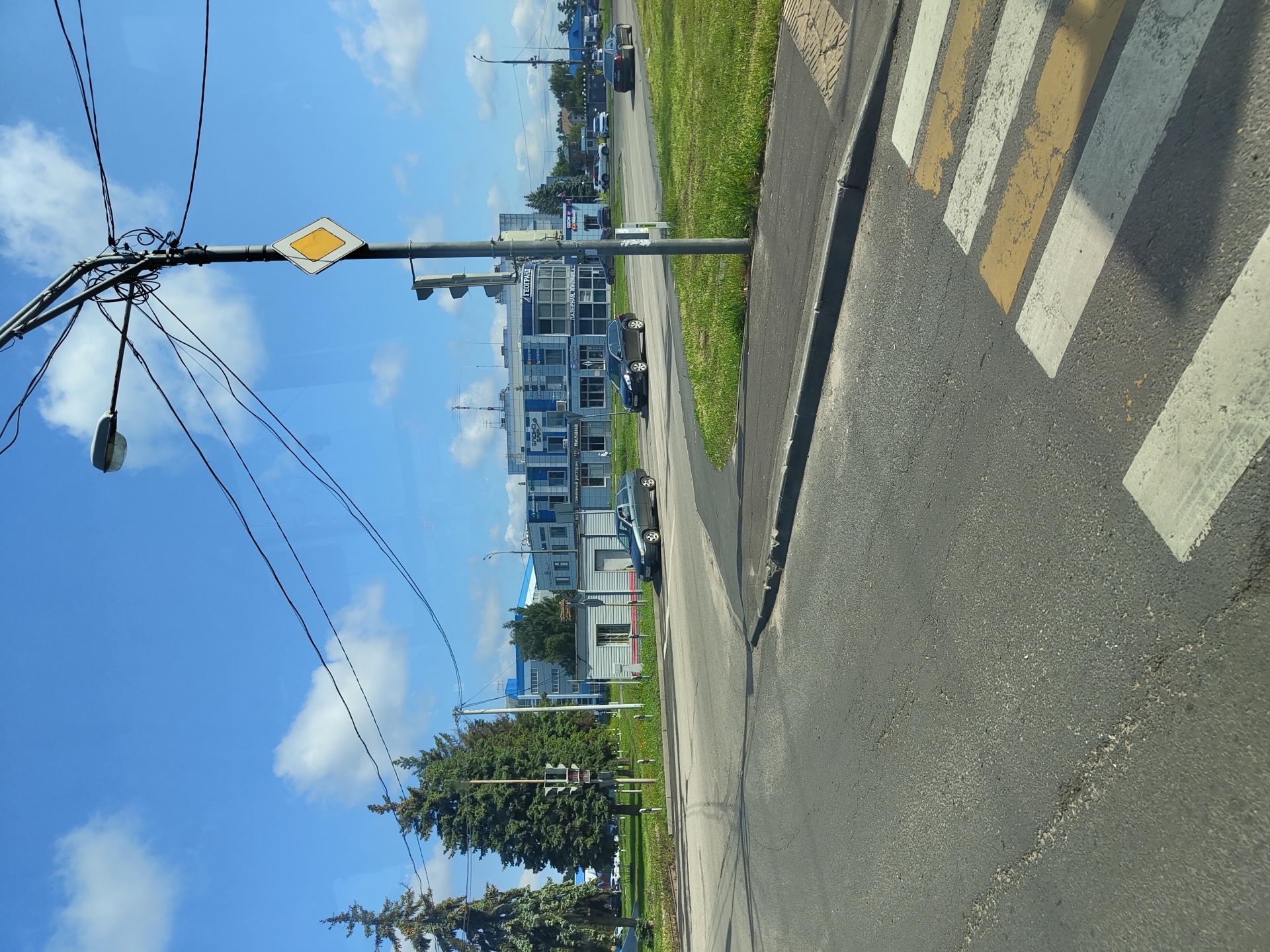 The image captures a street scene in the city of Zhukovsky, Russia, likely on a clear, sunny day. In the foreground, a pedestrian crossing with yellow and white stripes is visible on asphalt pavement. Beyond the crossing, a curved asphalt road leads to an intersection.
To the left of the intersection, a gray car is turning. Further back, several tall evergreen trees stand on a grassy median. Behind the trees, a low, gray building with a red stripe along its base is situated.
Dominating the middle ground is a modern, multi-story building with blue and white accents. One sign on this building reads "ГЕОГРАНЬ" (GEOGRANY). Another sign in front of it indicates "ОЗОН" (OZON) and "ЛАЗЕРНАЯ ЭПИЛЯЦИЯ" (LASER EPILATION). To the right of this building, a dark sedan is parked on the side of the road.
In the background, a streetlamp with multiple wires is prominent, and a diamond-shaped traffic sign with a yellow center is attached to its pole. Further in the distance, more buildings and trees are visible, along with other vehicles on the road. The sky is blue with scattered white clouds.