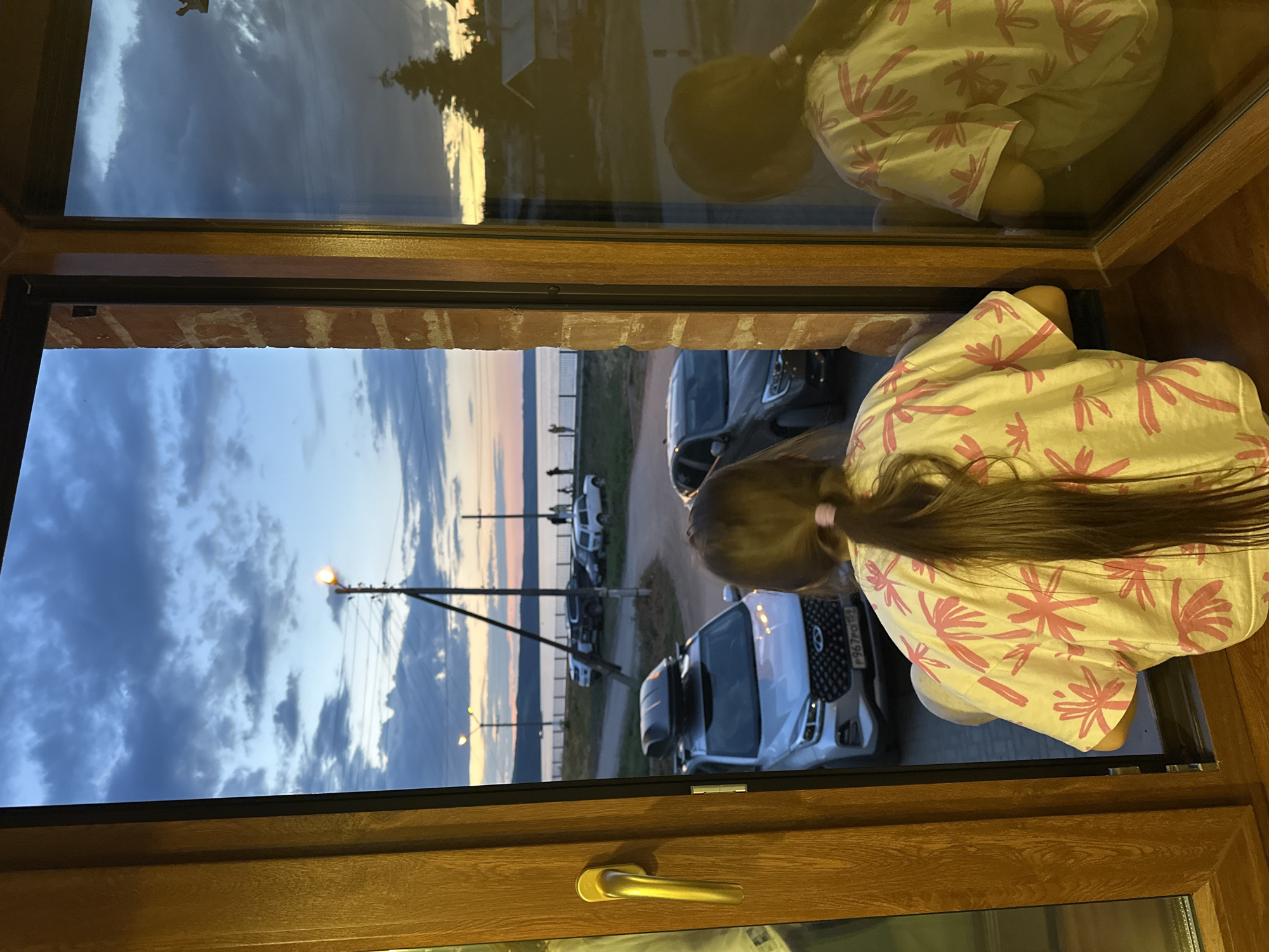 A young girl with long dark hair pulled into a ponytail is sitting on a windowsill, looking out of an open window at dusk. She is wearing a light-colored t-shirt with a pink floral pattern. Through the window, a body of water, likely the Volga River, is visible in the distance, with a shoreline dotted with buildings and trees. Several cars are parked along the road near the water. The sky is filled with dramatic clouds, transitioning from blue to shades of orange and yellow as the sun sets. The scene appears to be located in or near the city of Khvalynsk, Russia, given the geographical context. A reflection of the girl and the interior of the room can be seen on the glass of the adjacent window.