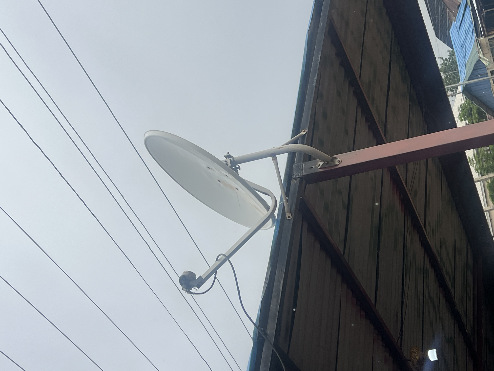 A satellite dish is mounted on a metal pole which is attached to the underside of a corrugated metal roof. The roof appears to be part of an outdoor shelter or awning. Multiple black electrical or communication wires are strung across the sky above the satellite dish and roof. The sky is overcast and a pale gray. In the background, a few buildings with blue and green roofs are visible, suggesting an urban or semi-urban environment. The scene is likely located in Jalingo, Nigeria, given the context. There are no people visible in the image.