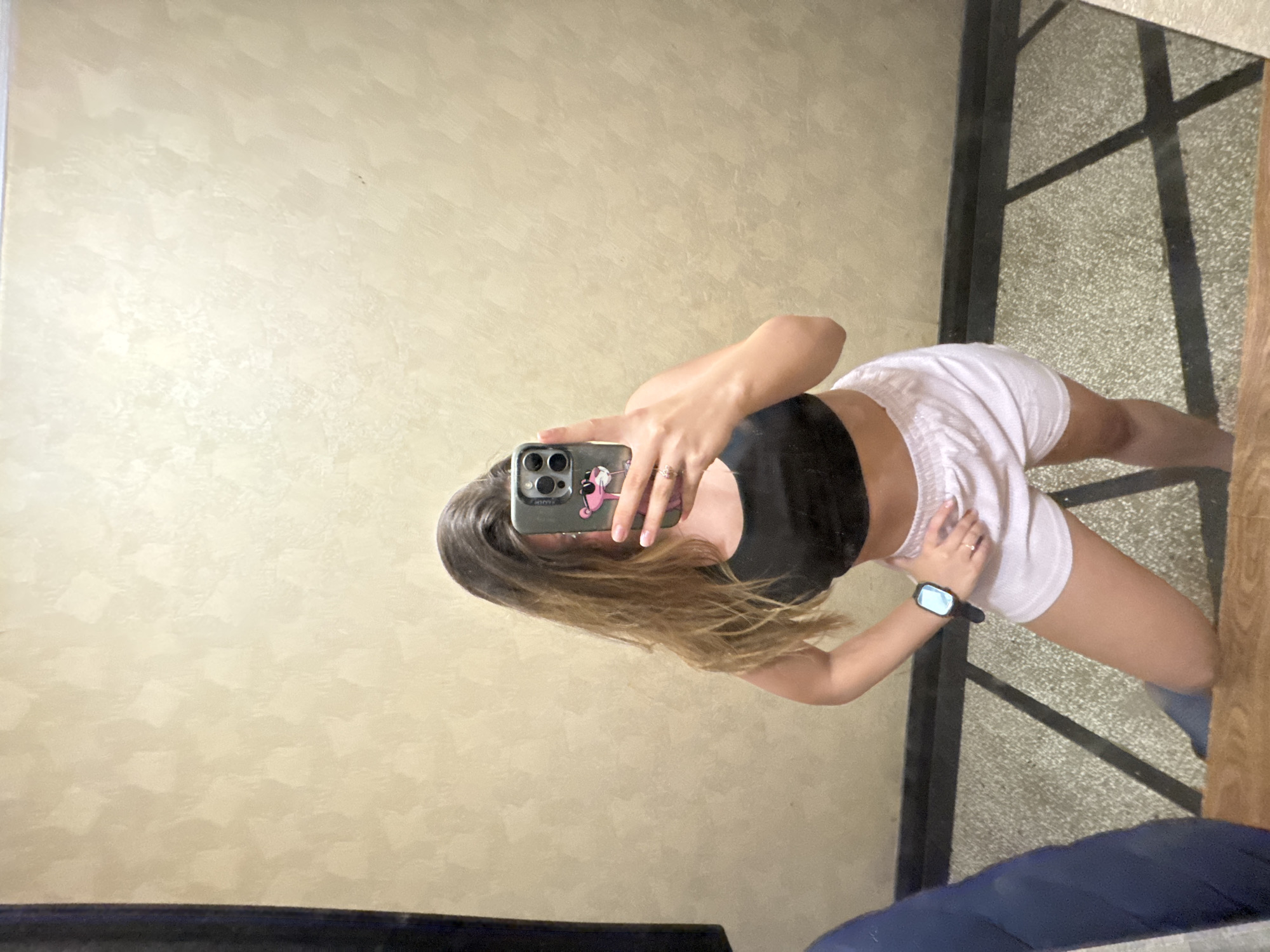 A full-length mirror selfie captures a person indoors, though the original image is rotated 90 degrees clockwise. The subject, a young individual with medium-length brown hair, is seen from the chest down, obscuring their face with a smartphone. They are wearing a black sports bra or crop top and white athletic shorts. A dark-colored smartwatch is visible on their left wrist, and a ring adorns their right hand.
The smartphone, a modern model with a triple camera array, has a distinctive grey-green case featuring a cartoon character, possibly the Pink Panther. The person is holding the phone up to take the picture, with part of their fingers visible on the screen.
The background reflected in the mirror reveals an interior setting. A light-colored wall with a textured, possibly brushed or stucco finish, displays subtle variations in shade and pattern. Below the waist, the reflection shows a light-colored, speckled surface, resembling concrete or terrazzo, crisscrossed by dark linear elements that could be part of a structural frame, window grid, or shadow patterns. Further into the reflection on the right, sections of a dark frame and a warm-toned wooden surface are visible, alongside a dark blue object, possibly upholstered furniture or a cushion, at the bottom.
The overall scene suggests daytime, given the soft, ambient lighting. While the image itself does not contain explicit geographical markers, the provided context indicates the location is Sofia, Bulgaria. No legible text is visible within the frame.