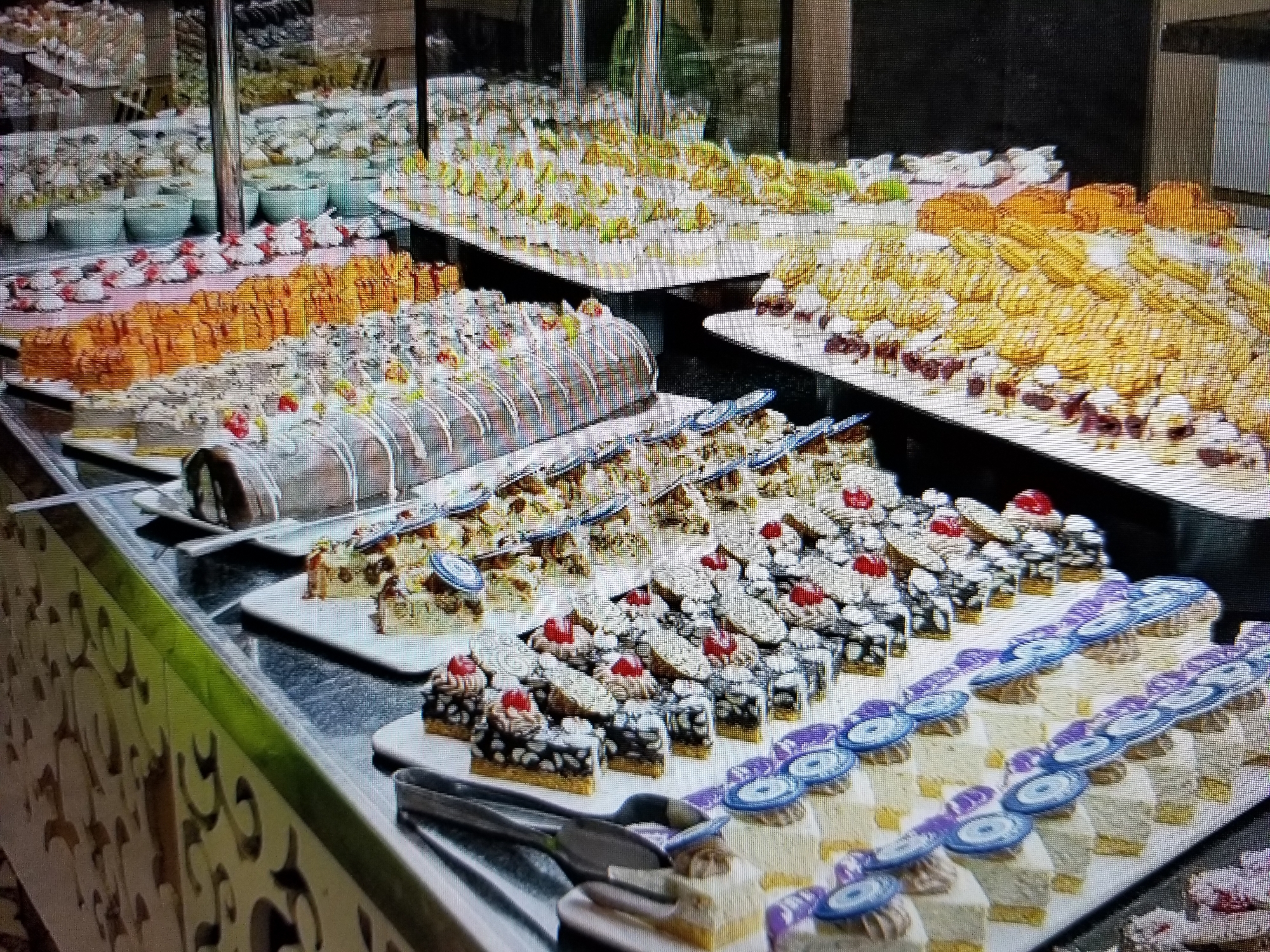 The image shows a buffet spread of various desserts. In the foreground, there are small square cakes topped with frosting and cherries, and behind them, rows of small pastries are arranged. To the left, a large rolled cake is visible. Further back, there are trays filled with various sweets, including small, round pastries. The desserts are displayed on white platters and are arranged on a dark countertop. In the background, more desserts are visible, along with some white bowls. The lighting appears to be artificial, and the overall scene suggests a dessert buffet, possibly in a hotel or restaurant. There are no people visible in the image. The location is Tiraspol, Moldova.