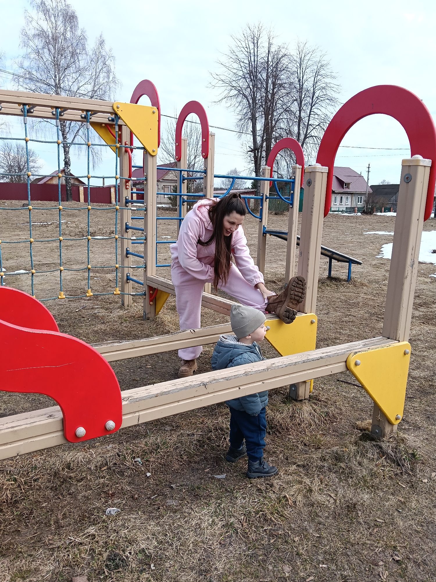 The image depicts a woman in a pink tracksuit and a young child at a playground in Красный Маяк, Russia. The playground features a structure with red and yellow accents, including a climbing net, ladders, and platforms. The woman is interacting with the child, who is wearing a grey hat and a blue jacket, near a platform on the structure where a boot is placed. The ground is covered with brown grass with some patches of snow. In the background, there are bare trees, houses with red roofs, and a cloudy sky.