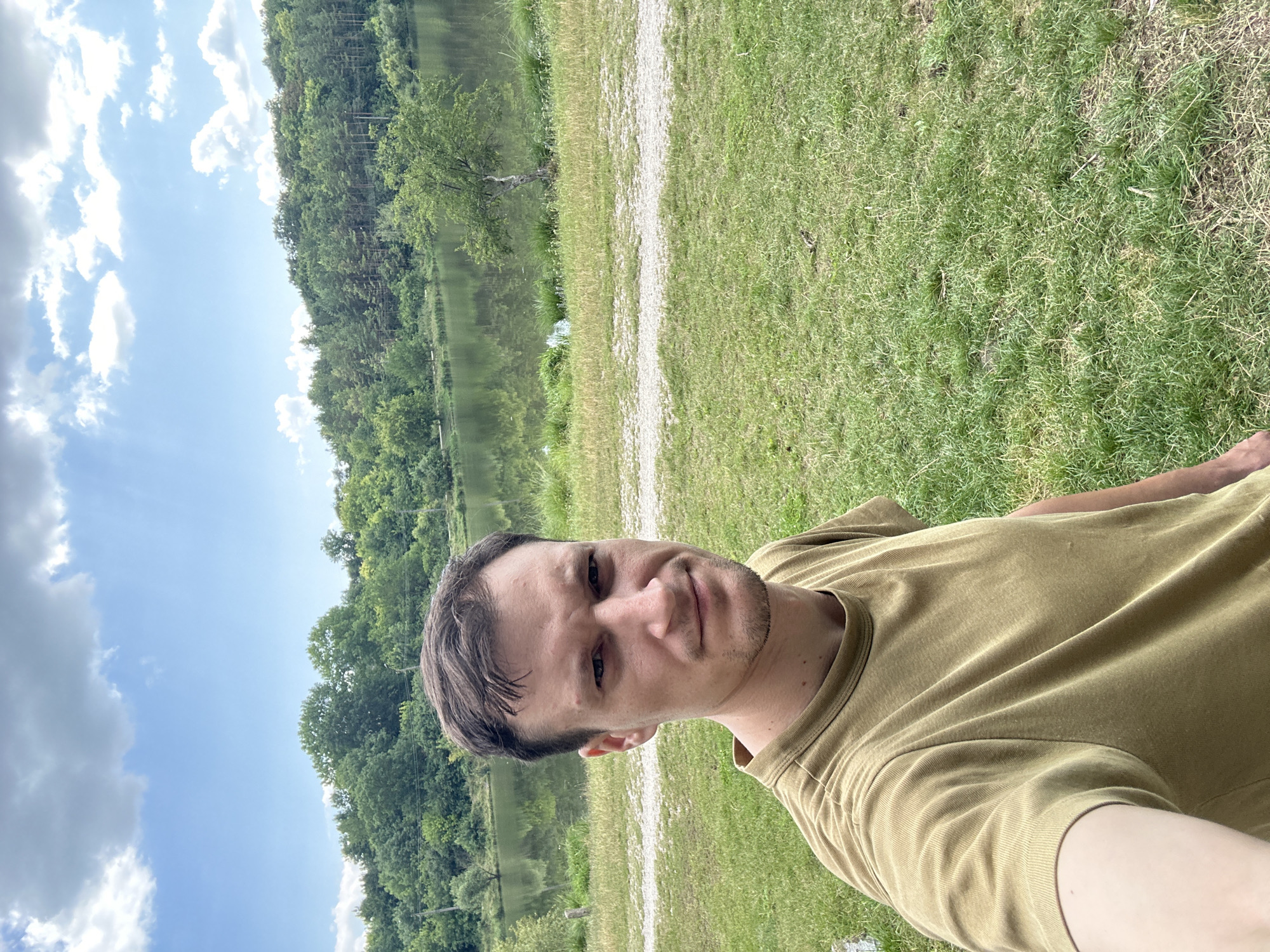 A man in a tan t-shirt takes a selfie in a grassy area near a lake. The lake is surrounded by a dense forest. The sky is blue with scattered clouds. The man is facing the camera and looking at it with a neutral expression. The background shows the natural landscape.