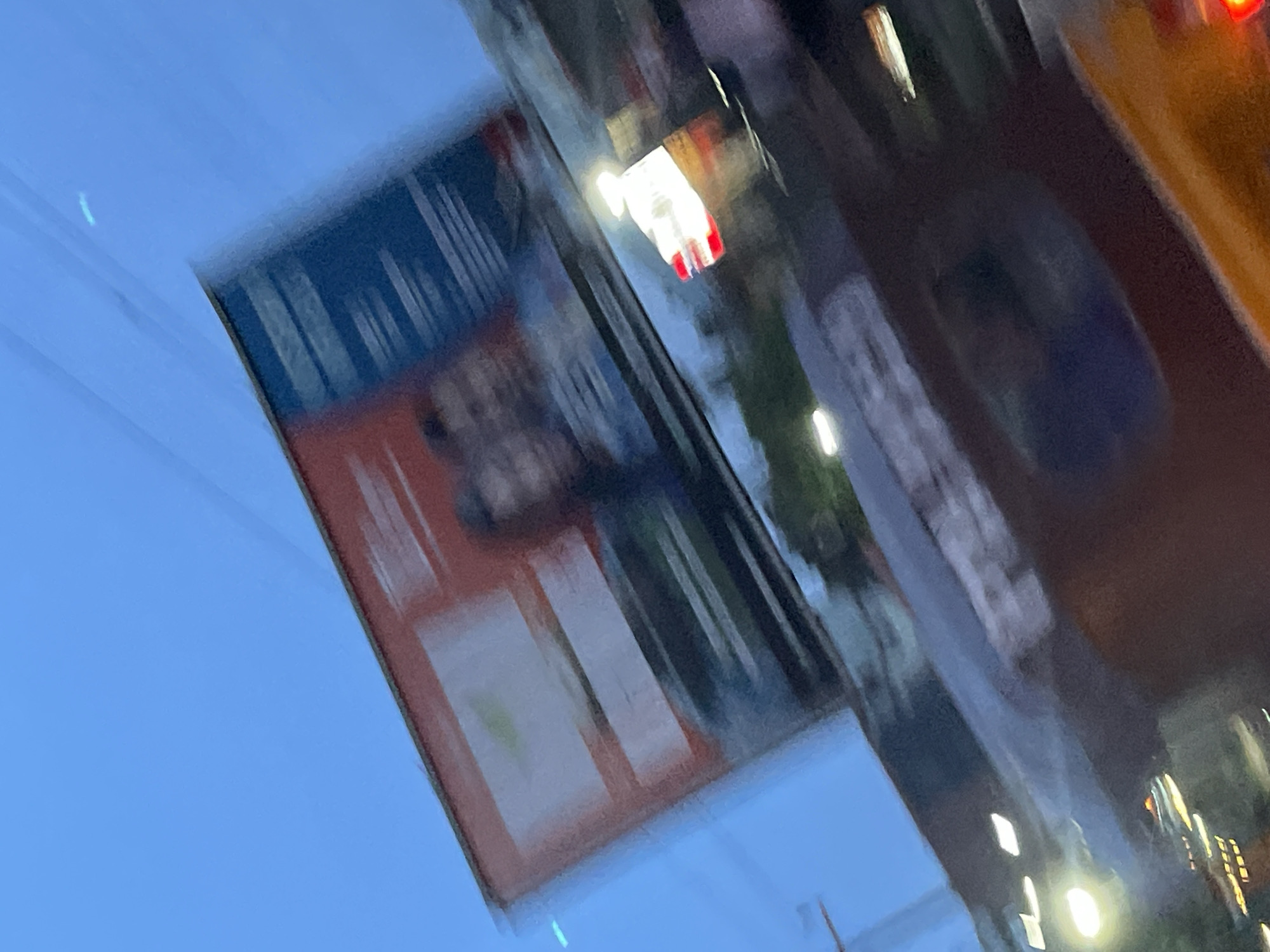 The image shows a blurry outdoor scene with a bright blue sky. A large billboard is visible at an angle, featuring a red and white design on the left and a dark blue section on the right with white text or graphics. The billboard appears to have images of two people, though their features are indistinct due to the blur. Below the billboard, a dark, reflective surface, likely the roof or hood of a vehicle, is visible. This surface reflects lights from below, appearing as streaks of white and yellow. One reflection shows a white light with red accents. The bottom of the image is dominated by a blurry, dark red or brown surface, possibly the interior of a vehicle or another object. The overall impression is of a fast-moving scene, possibly taken from a vehicle, at dusk or in low light conditions. The scene is identified as being in Jos, Nigeria.