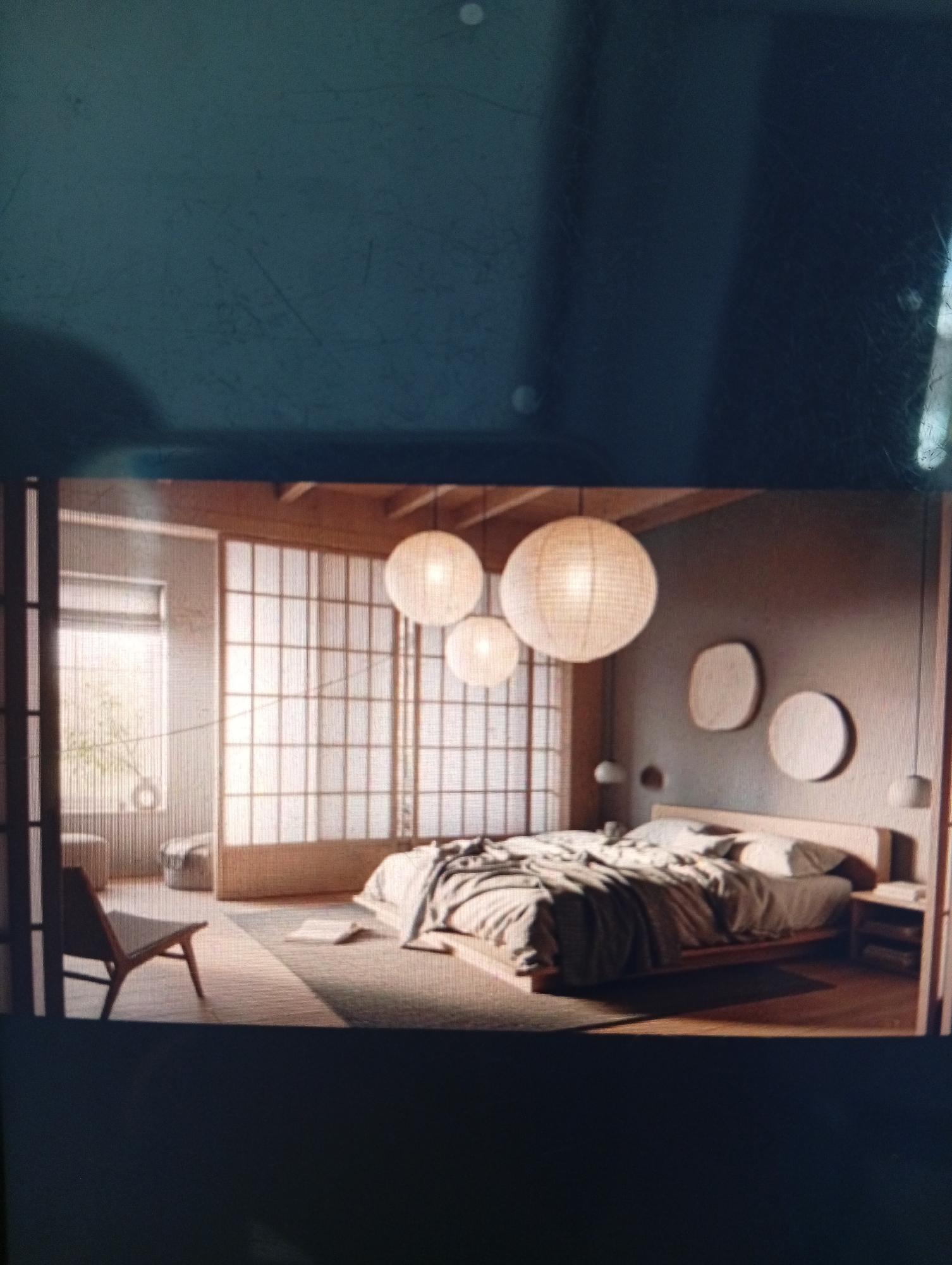 The image depicts a bedroom with a Japanese-inspired design. The room features a low platform bed with a greyish-brown duvet. Three paper lantern-style lamps hang from the ceiling, providing soft lighting. The walls are painted in a muted grey tone, adorned with three round, textured wall decorations. A shoji screen is present, acting as a partition. A wooden chair sits near the window, which is covered with a sheer curtain. A small bedside table is next to the bed. The overall aesthetic is minimalist and serene. There is no indication of a location in the city of Dutsen Alhaji, Nigeria.