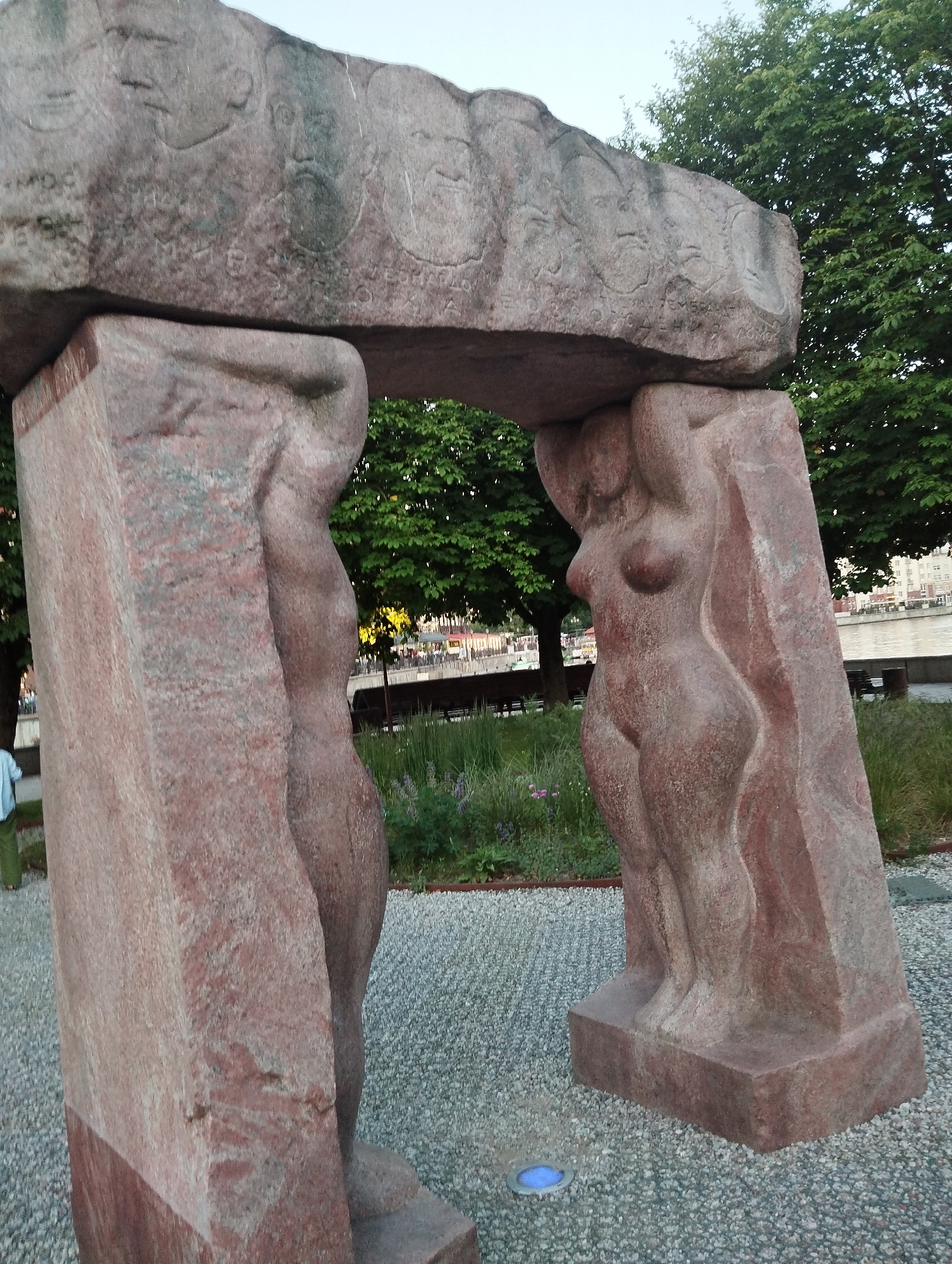 This is a medium shot of a large stone sculpture in Kaliningrad, Russia, with a river and buildings visible in the background. The sculpture consists of two large, carved figures supporting a massive stone lintel. The figures are stylized representations of human bodies, with prominent female anatomy. The lintel has carved reliefs of faces and inscriptions. The sculpture is made of reddish-pink granite. The ground around the sculpture is covered in small grey pebbles. In the background, there is lush greenery, a body of water, and several buildings. A person wearing a light blue shirt and green pants is partially visible on the far left. The sky is overcast. The overall scene depicts an outdoor public art installation.
