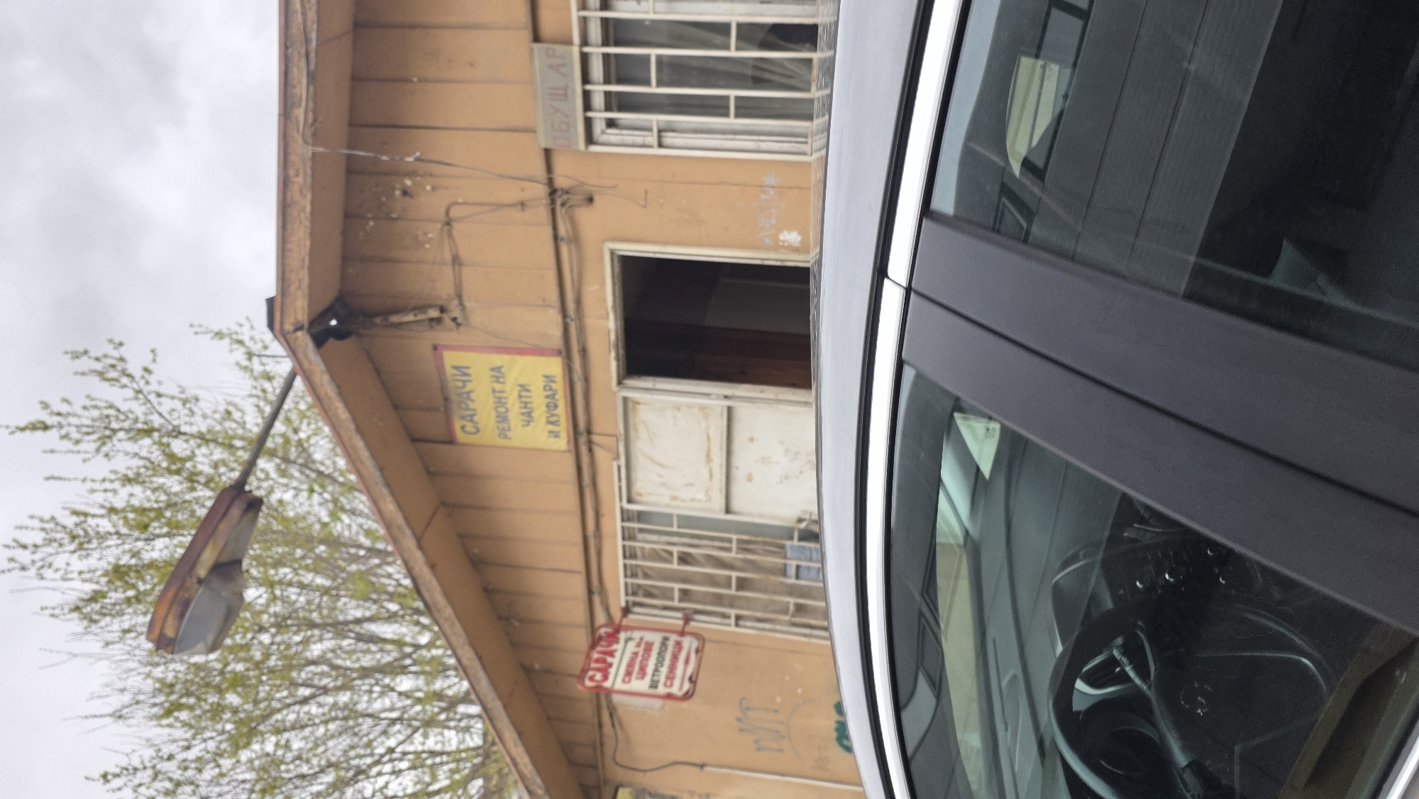 The image shows a building in Blagoevgrad, Bulgaria, with a sign that reads "САРАЧИ РЕМОНТ НА ЧАНТИ И КУФАРИ" in Bulgarian, indicating a repair shop for bags and suitcases. The building appears to be a single-story structure with a door and a barred window. Above the shop entrance, there is another sign with a word, but it is partially visible. A street lamp is positioned on the left side of the building. A car is parked near the building, partially obstructing the view. The sky above appears overcast.