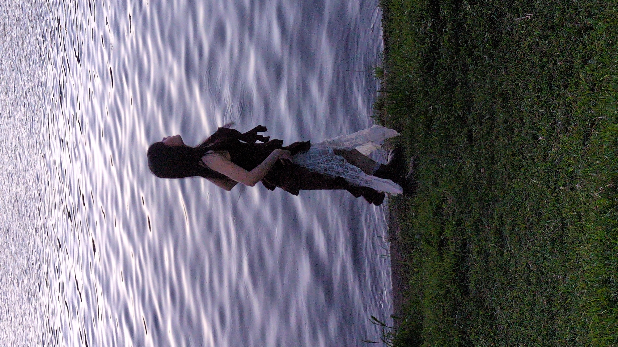 A woman stands on the grassy bank of a body of water. She is wearing a dark, layered dress with a white lace underlayer and dark boots. Her hair is dark and long, and she is looking up and to her right. The water behind her is rippled and reflects the sky. The image is taken in a natural setting, likely near a lake or river.