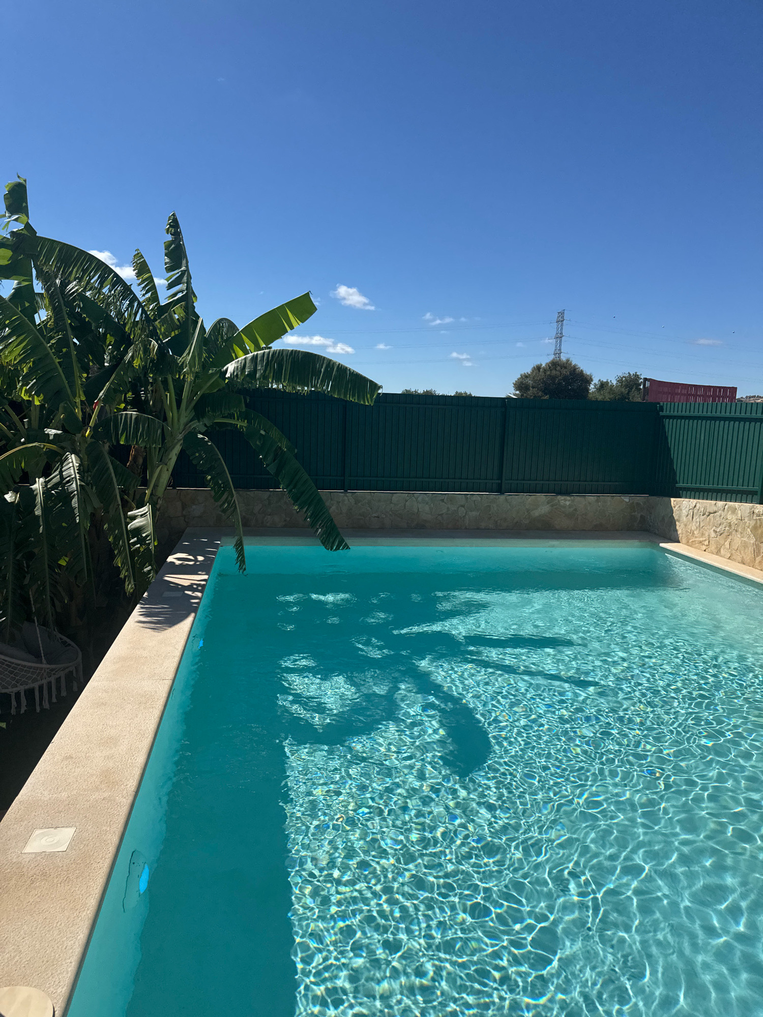 An outdoor swimming pool is shown under a clear blue sky with a few scattered white clouds. To the left of the pool, a large banana tree with broad leaves casts shadows onto the pool's surface. The pool is filled with clear, turquoise water, and the sun creates a rippling pattern of light on its surface. The pool's edge is bordered by light-colored concrete. Behind the pool is a stone wall and a tall, dark green metal fence. In the distance, behind the fence, a power line tower and some trees are visible, suggesting a location outside a densely populated urban area, possibly in the outskirts of Albufeira, Portugal. The scene is serene and evokes a sense of a warm, sunny day. There are no people or animals visible in the image.
