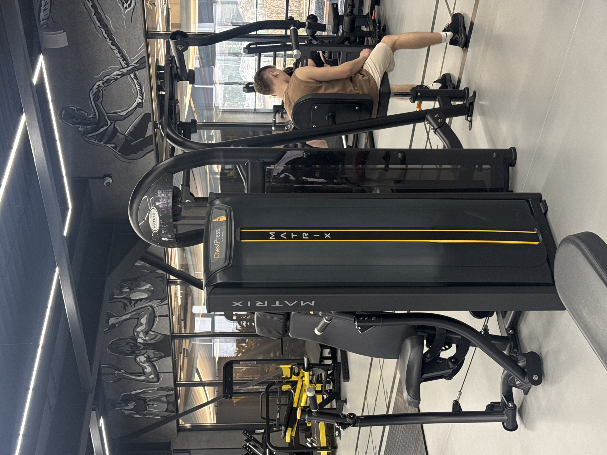 The image shows the interior of a gym. A man is sitting on a leg extension machine. The gym has workout equipment including a chest press machine by Matrix. The walls have murals of athletes lifting weights. The location is purportedly in Votkinsk, Russia.