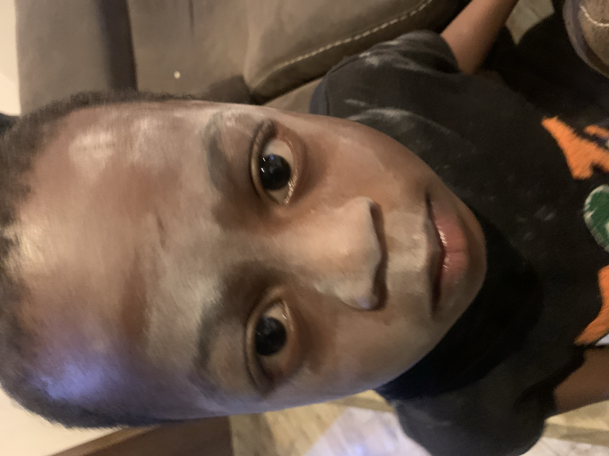 A close-up, slightly tilted, portrait photograph depicts a young person with brown skin. The person's face is partially covered with a white powdery substance. The person is wearing a dark-colored shirt, which also has spots of the white powder on it. The background shows a neutral-toned couch and part of a shoe. The location in the city of Jos, Nigeria, is not discernable from the image.