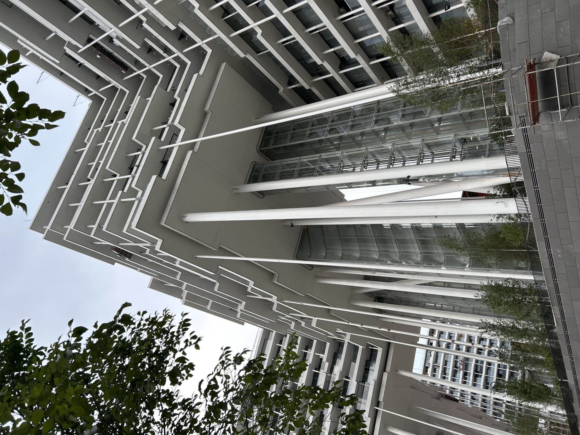 The image shows a modern white building with multiple levels and balconies, supported by numerous tall, slender columns. Two glass elevator shafts are visible between the columns. Trees and greenery are planted in front of the building. In the foreground, there is scaffolding and a stone-clad wall. The building appears to be located in Moscow, Russia, possibly part of the Moscow City business district, given its contemporary architecture. The sky is overcast. There are no visible people or discernible actions.