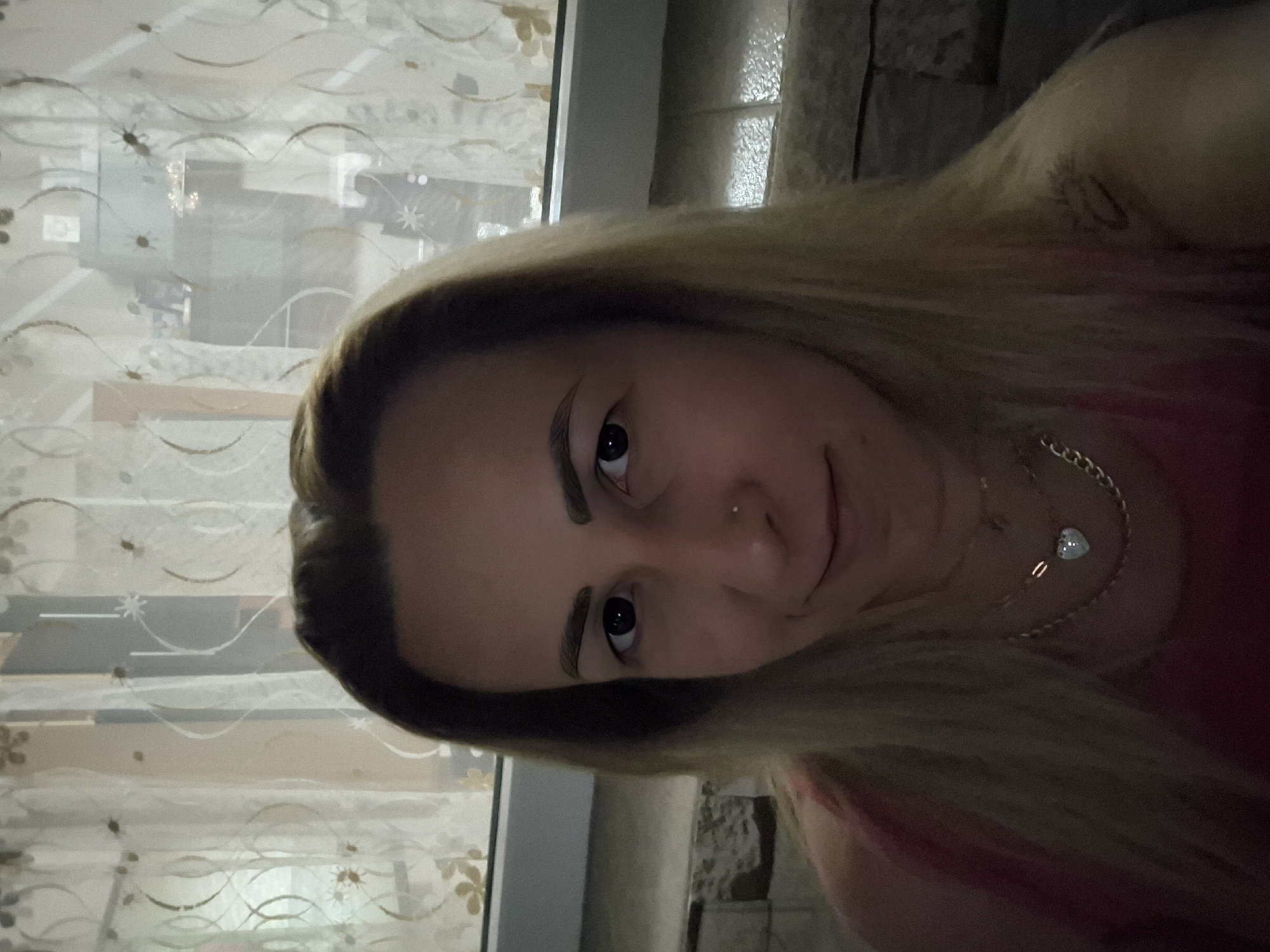 A medium shot selfie of a woman with long, blonde hair and light skin, looking into the camera. She is wearing a pink top and multiple necklaces, including one with a heart-shaped pendant. She has a nose ring and appears to have a tattoo on her right shoulder. The background shows a window with sheer curtains and some furniture. The scene is indoors, with the lighting suggesting it is daytime. The woman's expression is neutral, with a slight smile.
