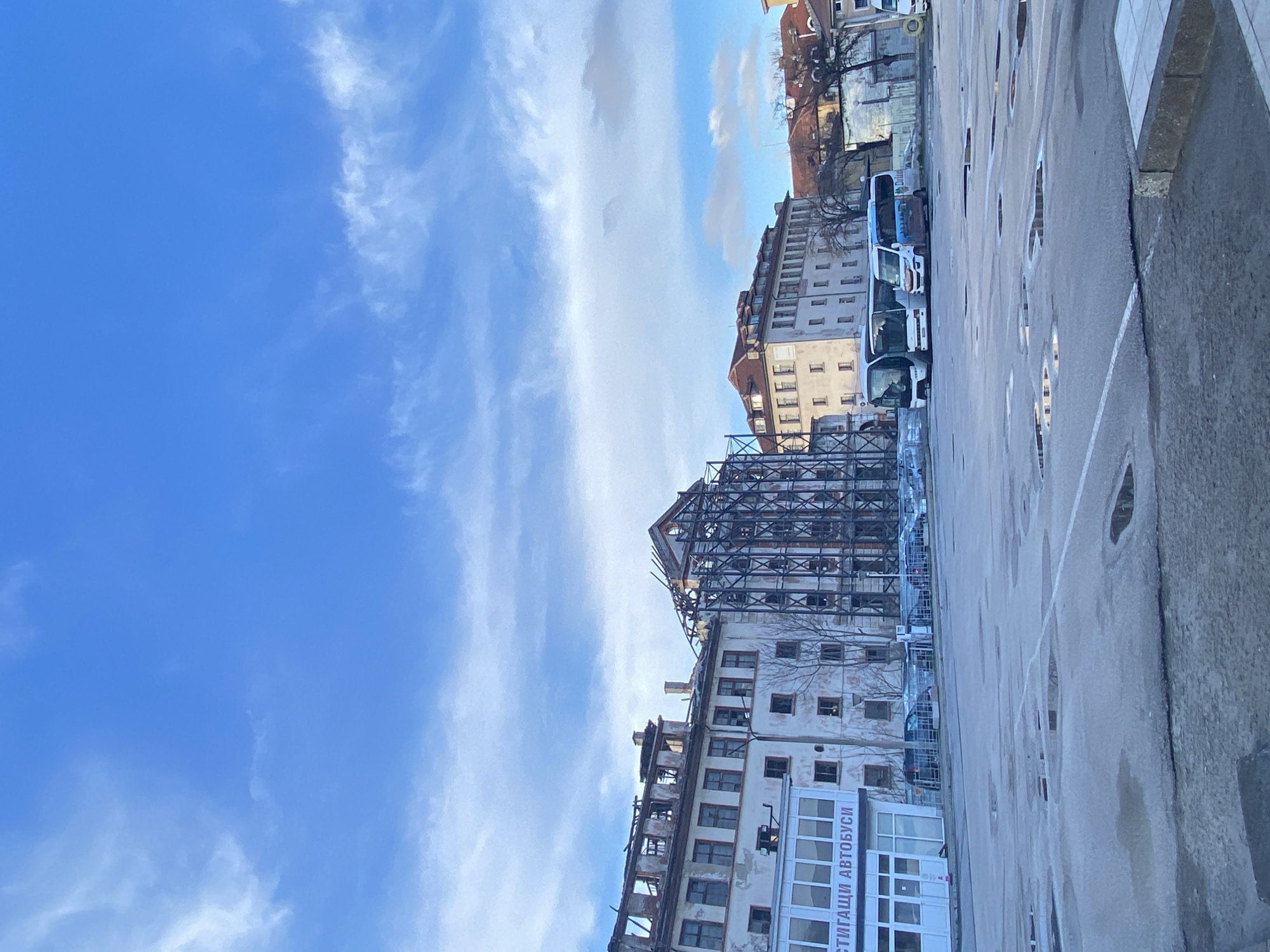 The image depicts an urban scene in Plovdiv, Bulgaria. In the foreground is a concrete ground or street surface that is wet, possibly from rain. On the right side of the image is a curb or sidewalk. Looking up, there are several buildings with varying architecture. One appears to be undergoing renovation or construction, with scaffolding surrounding its upper floors. Further in the background are other buildings, some with visible trees. Several white buses are parked alongside the buildings. The sky is predominantly blue with streaky white clouds. The overall scene suggests a city street with a construction site and parked vehicles under a partly cloudy sky.