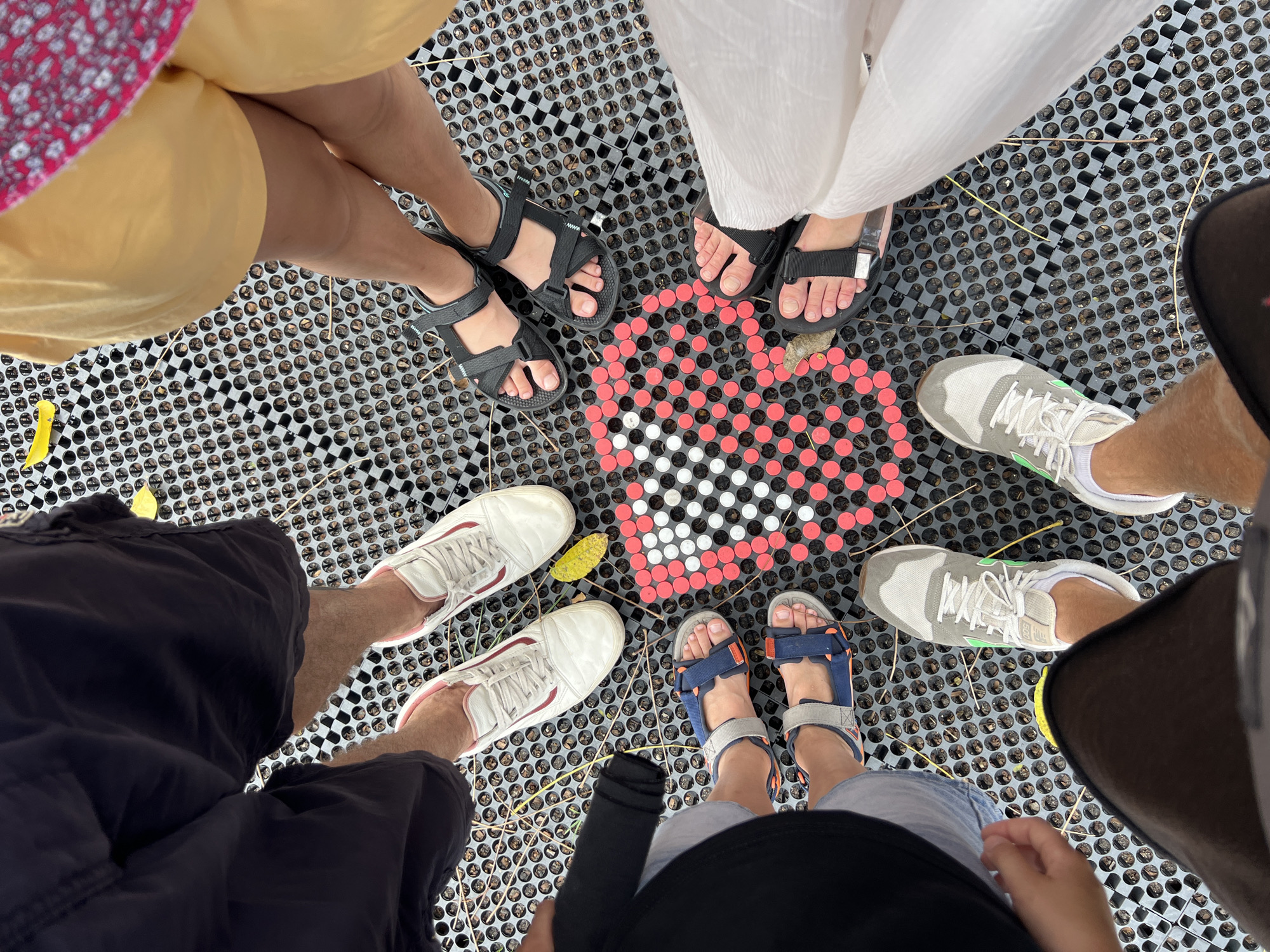 An aerial view shows seven pairs of feet standing around a heart-shaped mosaic made of red and white circular objects on a textured grey surface with circular perforations. The individuals are wearing various footwear including white sneakers with pink accents, blue and orange strap sandals, black strap sandals, and grey sneakers with green accents. Some individuals are wearing shorts, while others are wearing skirts or dresses. The heart mosaic is formed by approximately 300 small circular objects arranged in a heart shape with a black border. The background surface appears to be a metal grate or similar material with a repeating pattern of circles. The scene is outdoors and the lighting suggests daytime. There is no specific indication of the city of Kediri, Indonesia in the image.