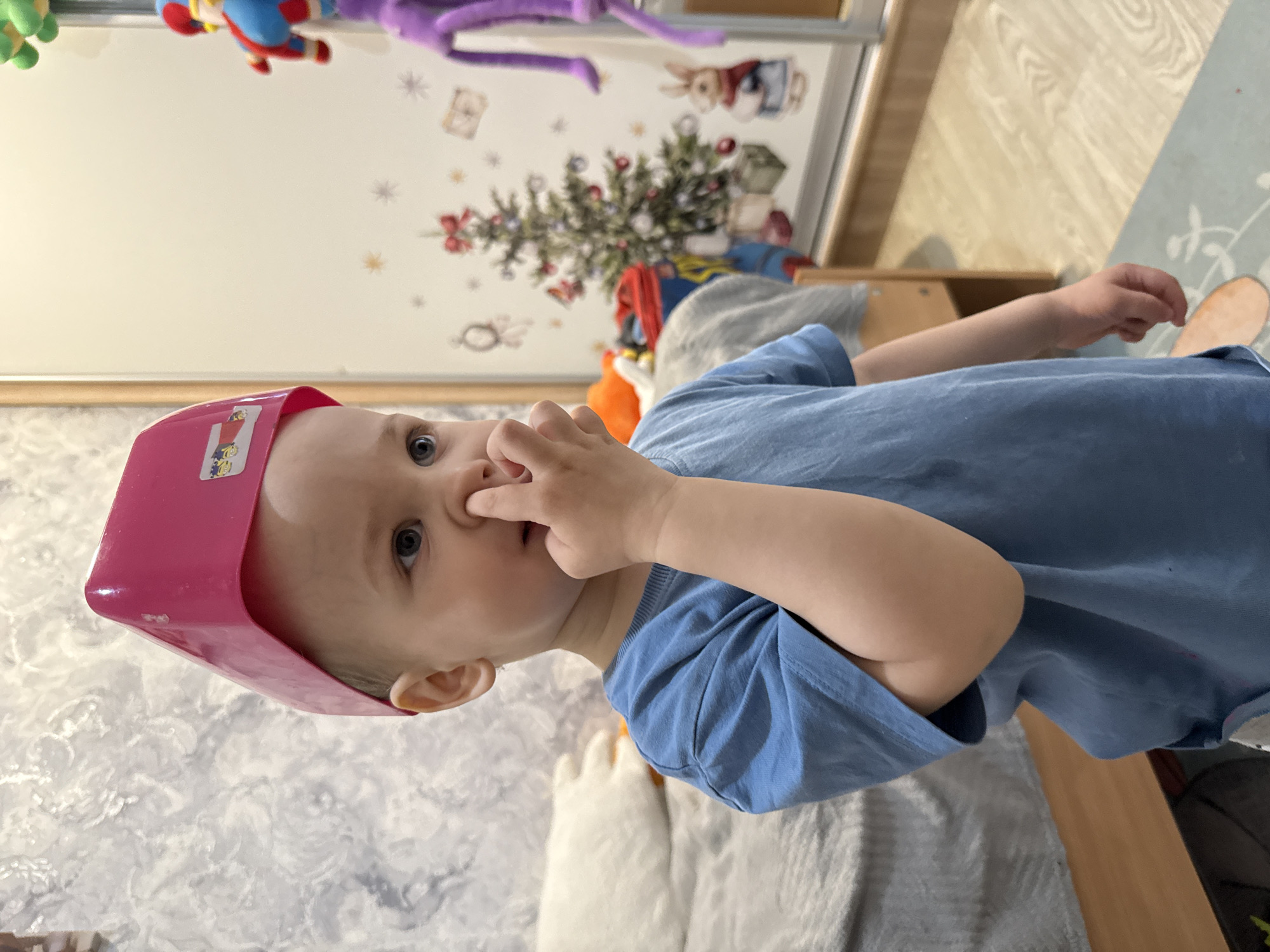 A close-up shows a small child with fair skin, wearing a bright pink plastic bucket on their head. The child is wearing a blue t-shirt and has their fingers inserted into their nose. The background includes a cabinet or wall with cartoonish decorations, including a Christmas tree and hanging plush toys. The environment appears to be indoors, possibly in a child's bedroom or play area. There is no visible location in the city of Unknown, Unknown.