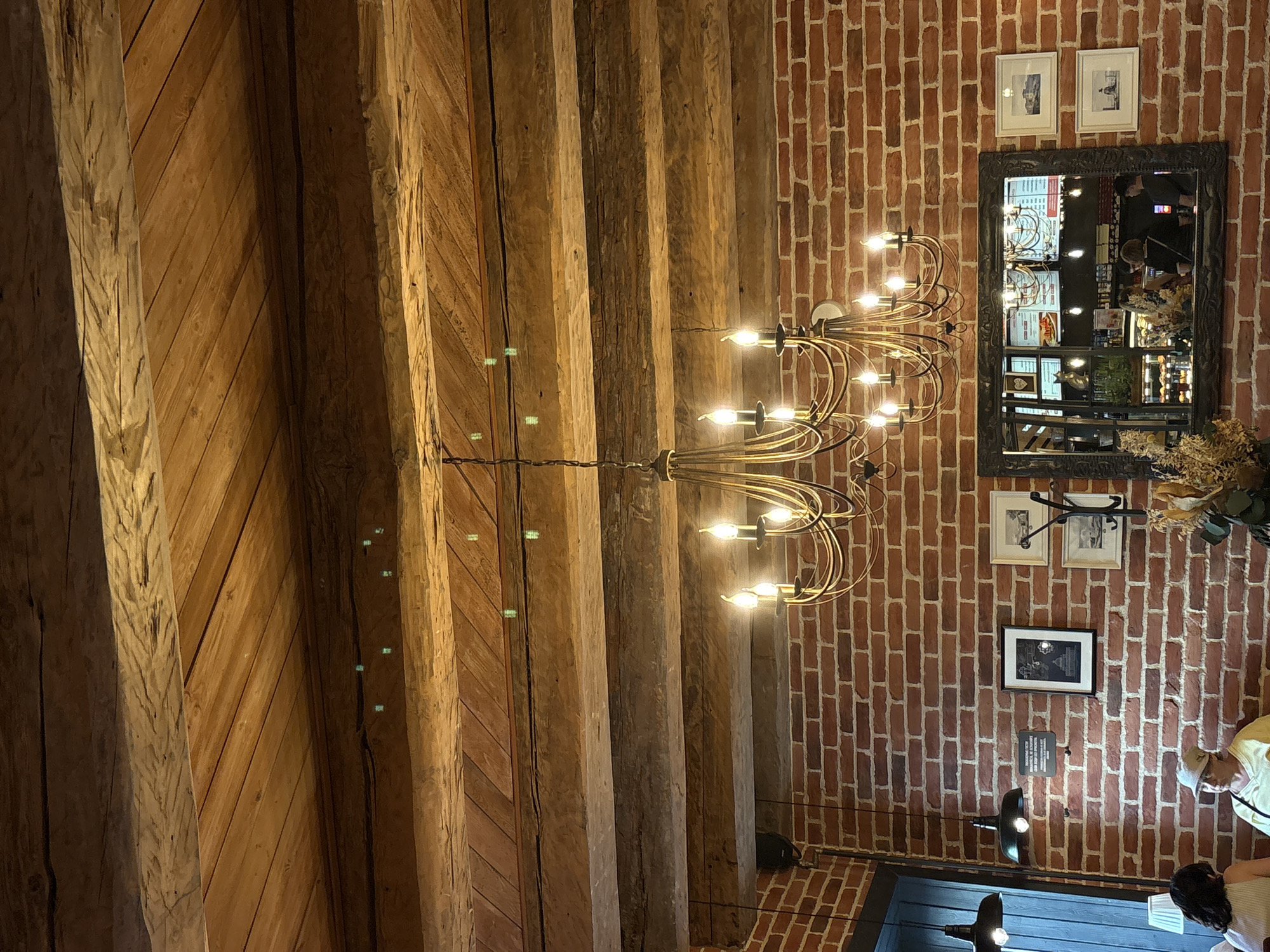 The image depicts the interior of a commercial establishment, likely a cafe or restaurant, in Saint Petersburg, Russia. The ceiling is constructed from parallel, rough-hewn wooden beams interspersed with lighter-toned wooden planks, some laid diagonally. Small, faint, greenish reflections are visible on various surfaces of the wooden ceiling elements. A prominent, two-tiered, gold-toned chandelier with multiple illuminated candelabra-style bulbs hangs centrally from the ceiling.
The walls are composed of exposed red-brown brick with white mortar. On the right wall, a large, dark-framed mirror reflects a service area behind the viewer, showing a counter, menu boards with legible text and graphics, and approximately five indistinct individuals. To the right of the mirror, three smaller framed monochrome prints, likely landscapes or architectural scenes, are mounted; one is positioned above two stacked vertically. A dark, wall-mounted coat rack is installed between the upper and lower right pictures. On the mid-left brick wall, a dark plaque with white text and a small, dark-framed picture are visible. A dark blue or black vertical panel, possibly part of a door or window frame, is visible on the far left.
Two black pendant lamps hang from the ceiling on the left side of the room. In the lower-left foreground, two individuals are seated. One has dark hair, partially visible. The other, wearing a light yellow top and a light-colored brimmed hat, faces towards the right. A dried floral arrangement is present in the lower-right foreground. The overall lighting is warm, emanating primarily from the artificial fixtures.
