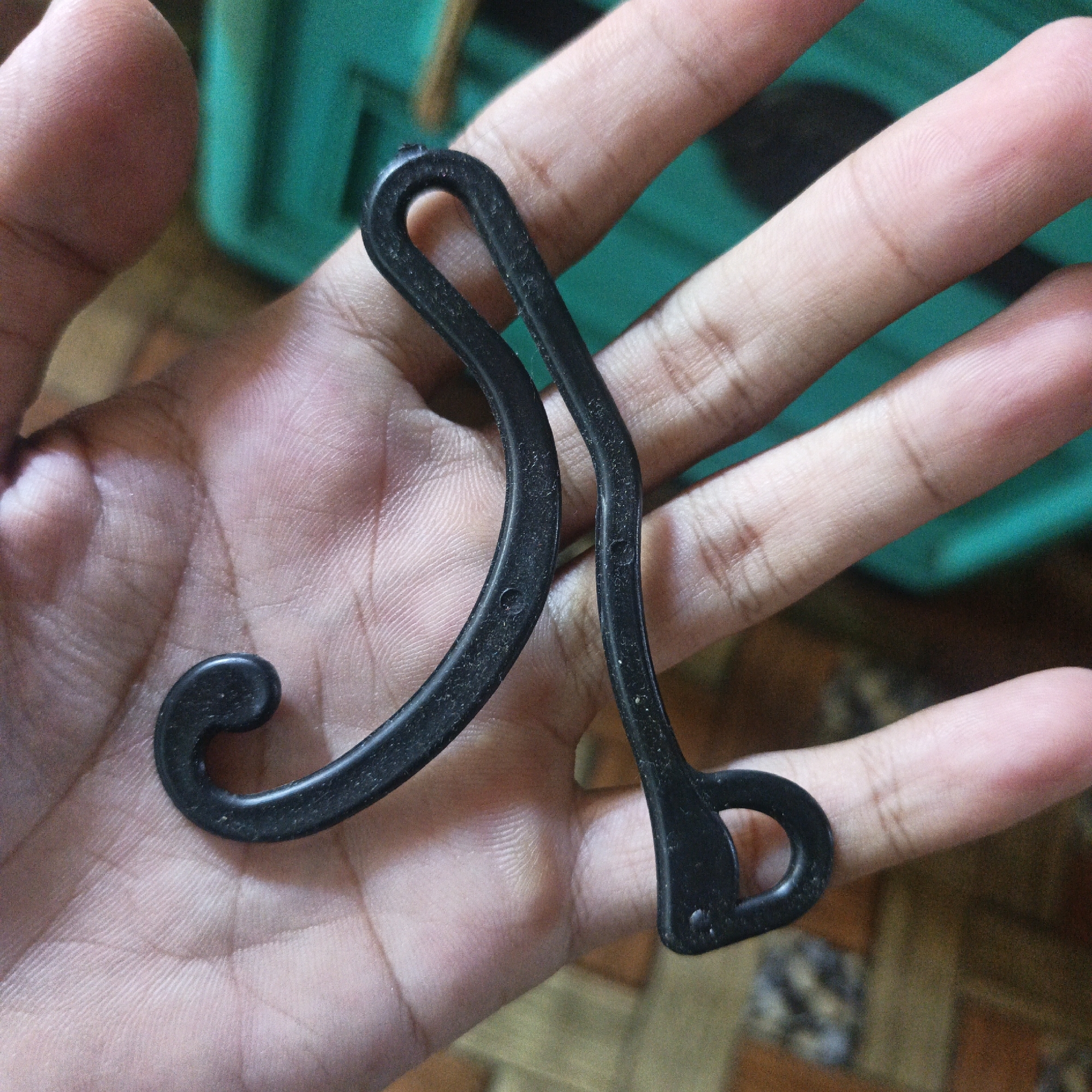 A close-up shot captures a hand holding two black plastic earbud hooks. The hooks are curved and one has a small curl at the end. The skin of the hand shows visible fingerprints and lines. In the background, a blurred teal object and a wooden floor are visible. The lighting suggests an indoor setting. There is no indication of the location being in Sungai Rasau, Indonesia.