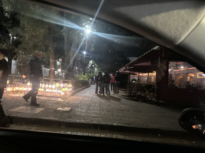 The image depicts an evening scene in a public square or park in Mexico City, likely in the Coyoacán borough near Jardín Centenario or Plaza Hidalgo. The view is from inside a vehicle, with portions of the dashboard or windshield frame visible at the top and bottom.
In the foreground, on the left, a male in dark clothing walks right-to-left across a paved area. Adjacent to him is a street vendor's stall displaying multiple small, colorful, illuminated lanterns or decorative lights arranged in a grid-like structure. A person is seated behind this stall. The paved pedestrian path extends into the midground, where several individuals walk or stand, including a small group centered in the frame. The area is illuminated by a prominent streetlamp overhead, casting a bright flare. Tall trees with dark foliage line the background of the path.
On the right side of the midground, a single-story building with a dark red tiled roof and a covered outdoor seating area is visible. The outdoor section contains tables and chairs, some occupied by patrons. The building's interior is lit, showing reflections in windows. A cart or kiosk-like structure is positioned near the restaurant's entrance. The overall environment is characterized by active pedestrian traffic, street vending, and a restaurant with outdoor dining.