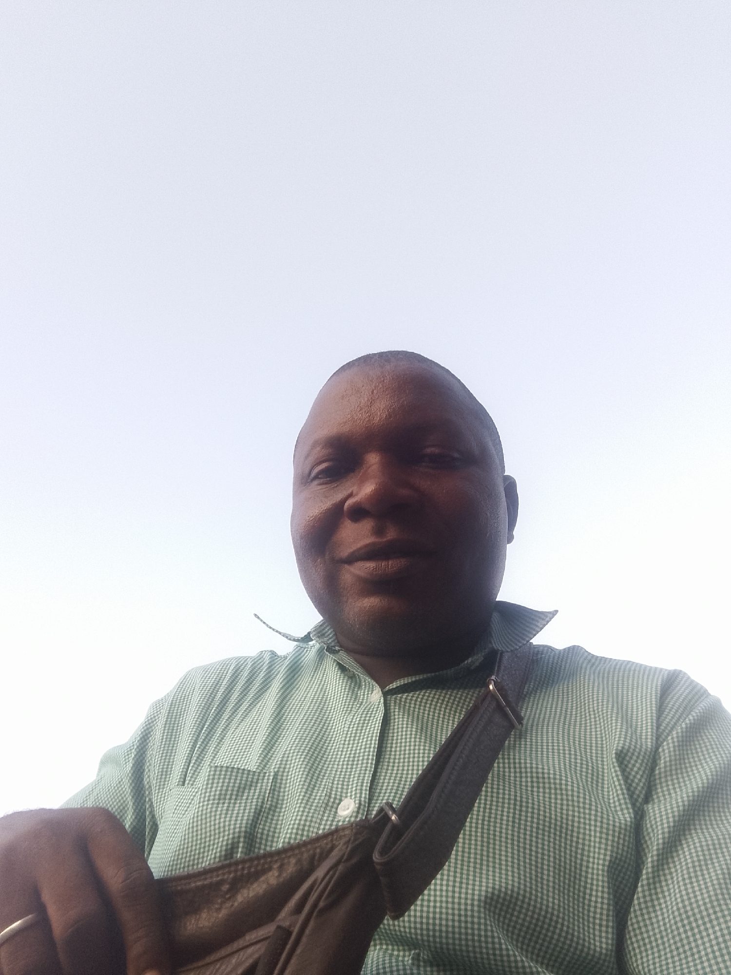 A medium shot shows a dark-skinned man wearing a light green checked shirt and a dark-colored bag with a strap. He has a ring on his finger. The background is a blurred white. The location is Lokoja, Nigeria.