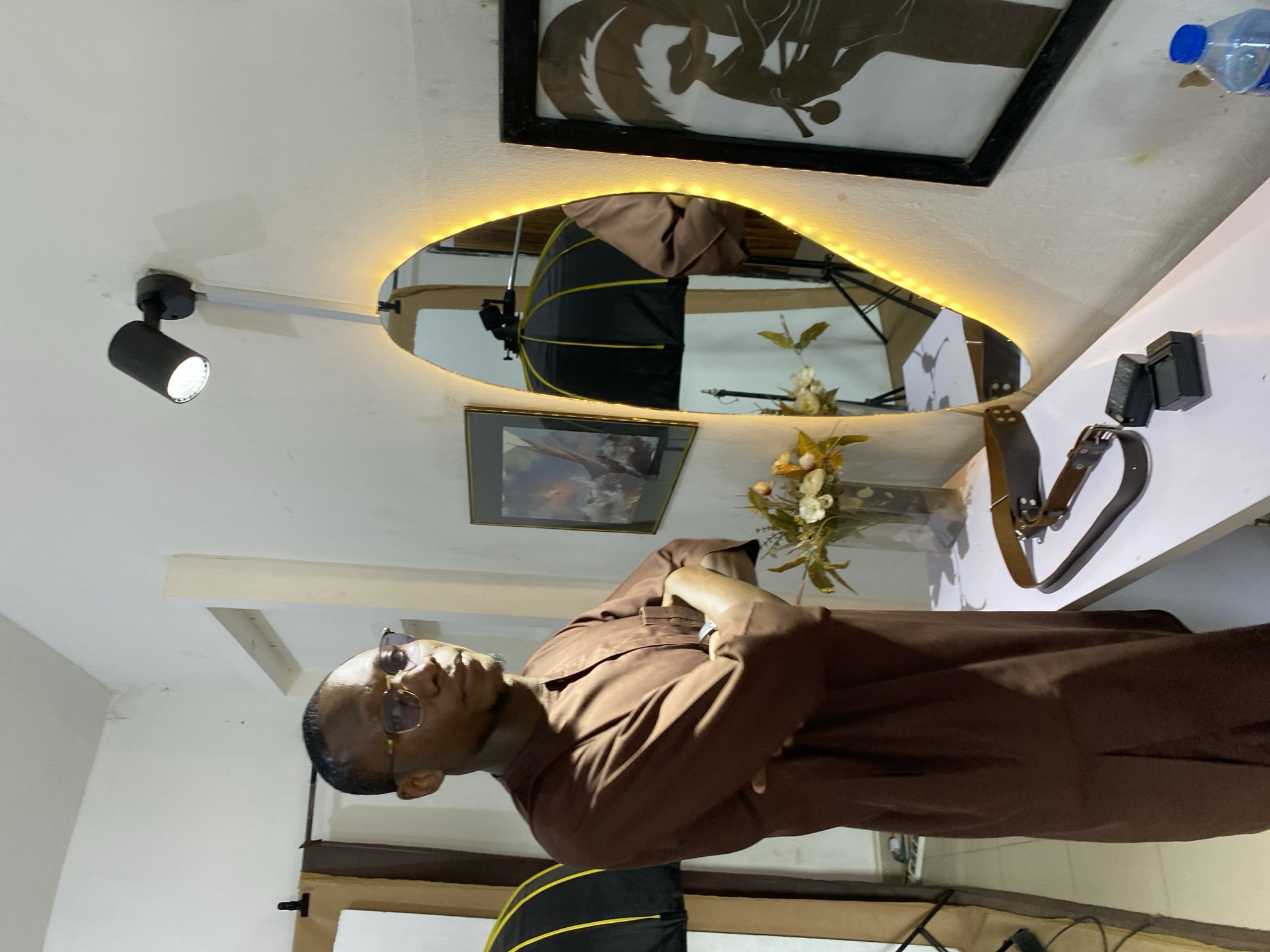 An adult male stands indoors, facing the viewer with his arms crossed. He is wearing a brown long-sleeved tunic and dark sunglasses. His short dark hair and facial hair are visible.
The setting appears to be a well-lit indoor space, possibly a photography studio or a versatile creative office. The walls and ceiling are a light color. To the left of the man, a black spotlight is mounted on the ceiling or upper wall, directing light into the space. Faint discoloration or marks are visible on the ceiling.
Behind the man, a prominent feature is an irregularly shaped mirror framed with an illuminated LED strip, casting a warm glow. The mirror reflects a black and yellow photography softbox or umbrella and its stand, indicating a photography setup. Above the mirror, a framed piece of art with a dark silhouette design hangs on the wall. Below the mirror, another smaller framed picture is visible, alongside a vase holding artificial flowers and foliage.
To the right of the mirror, on a white table or shelf, are several objects: black electronic devices (possibly chargers or battery packs), a dark leather belt with metal buckles, and a clear plastic water bottle with a blue cap. Partially visible in the lower foreground, to the man's left, is another section of a black and yellow photography softbox. The man's expression is neutral, and he appears to be posing for the photograph. There is no visible text in the image. The location context provided is Bukuru, Nigeria.