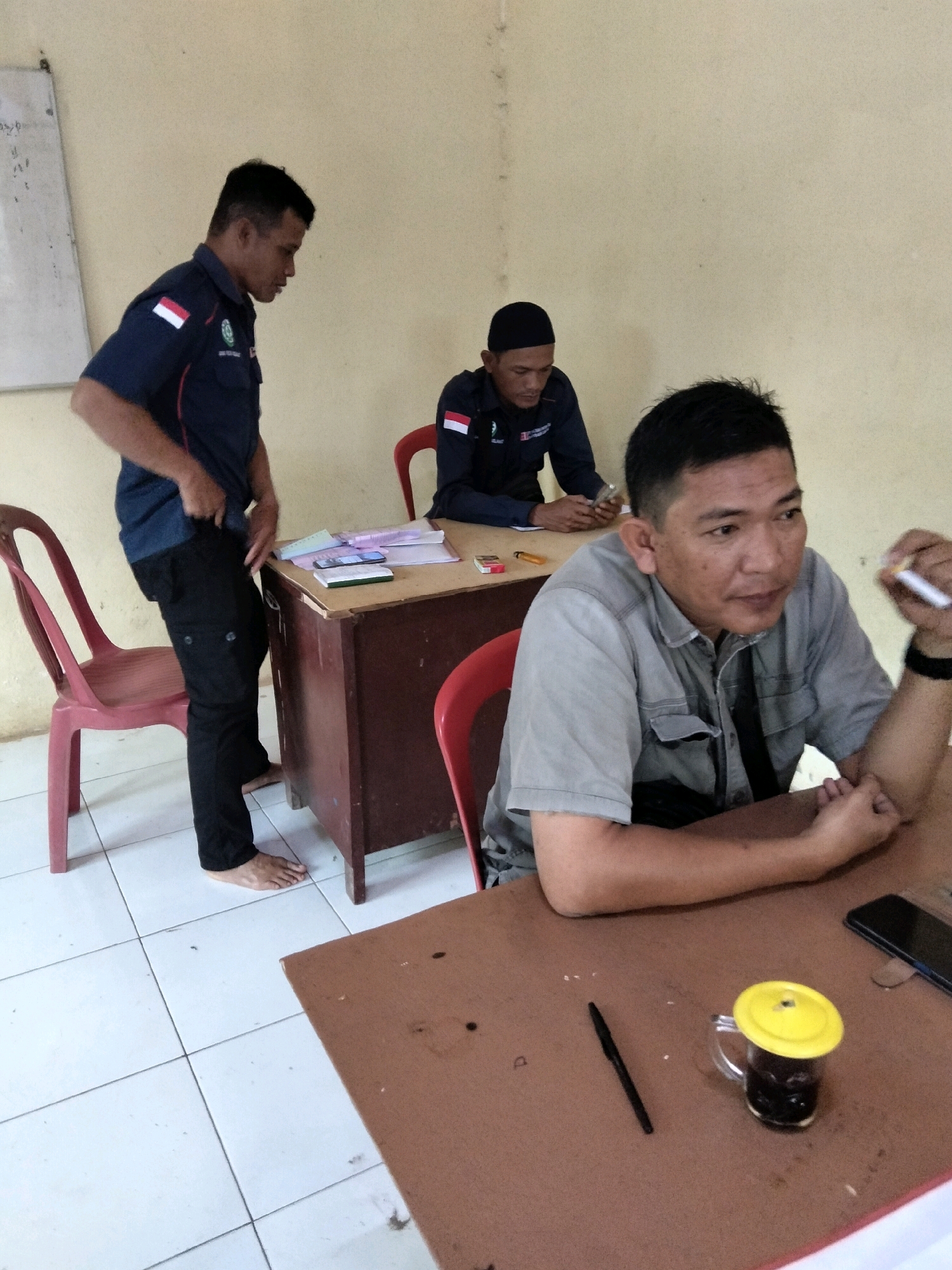 In a room with beige walls and white tiled floors, three men are present. The man on the left, wearing a dark blue shirt with red and white insignia on the sleeves and chest, stands next to a wooden desk. He has short black hair and is wearing black pants. To his right, behind the desk, a man in a dark blue uniform with a black cap is seated. He has short dark hair. In the foreground, a third man with short dark hair is seated at a table, wearing a grey collared shirt. He is holding a white pen in his right hand and appears to be looking to his right. On the table in front of him are a glass of dark liquid with a yellow lid, a black pen, and a smartphone. The desk behind the man in the uniform has papers, a ruler, and a mobile phone on it. Two red plastic chairs are visible in the image. The scene suggests an indoor office or meeting space in Cinta Manis Lama, Indonesia.