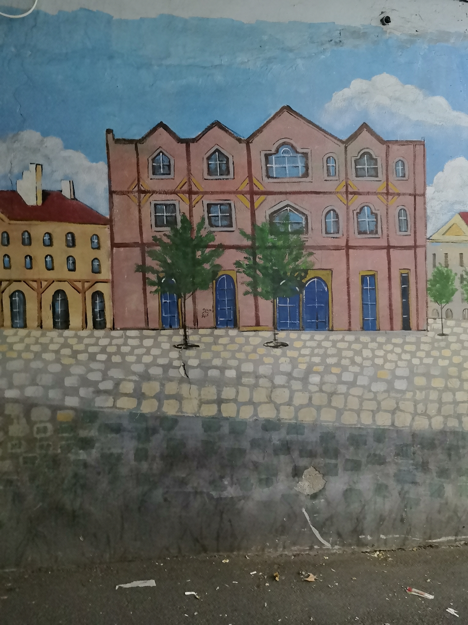 This is a mural painted on a wall depicting a row of buildings with a cobblestone street in front. The buildings are rendered in a stylized, somewhat naive manner. The building on the left is yellow with a red roof and numerous small, arched windows. The central building is larger, with a pink facade, arched windows, and what appear to be blue double doors. Two small, green trees are painted in front of this building. To the right, another building with a yellow roof is partially visible. The sky above is painted blue with white clouds. Below the buildings, a section of gray wall with a crack is visible, followed by a dark, ground-level area with debris. The mural has signs of wear and tear, with peeling paint and cracking in the wall. It is difficult to pinpoint a specific location in Yerevan, Armenia, based solely on the artwork itself.