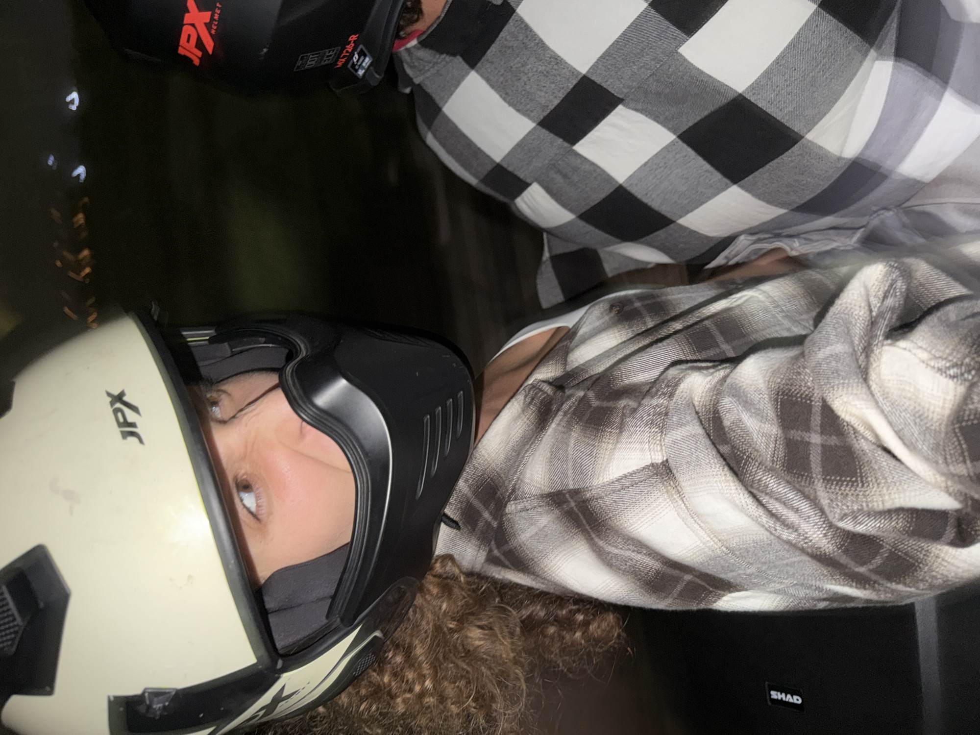 An overhead, low-light photograph, taken at night in North Kuta, Indonesia, captures two individuals, presumably on a motorcycle or scooter. The image is oriented vertically but depicts the subjects as if viewed from above, looking down.
In the foreground, a person is seen wearing a light-colored (off-white or cream) full-face motorcycle helmet, featuring the brand "JPX" prominently on the side. Their eyes are visible through the visor opening, looking towards the right, with light brown irises. Curly brown hair spills out from beneath the helmet. This individual wears a plaid flannel shirt in shades of grey, white, and dark grey.
Behind and slightly to the left of the foreground subject, another person is partially visible, wearing a black full-face helmet with "JPX HELMET" and "MX720-R" written on it. They are dressed in a distinct black and white checkered plaid shirt.
The background is dark and blurred, with faint, out-of-focus light sources suggesting artificial lighting from a nighttime setting. Below and to the right of the foreground person, a black luggage box or compartment is visible, with the brand name "SHAD" in white letters. The overall scene suggests the individuals are either preparing for a ride, riding, or have just finished an outing on a motorbike, common in the given location.