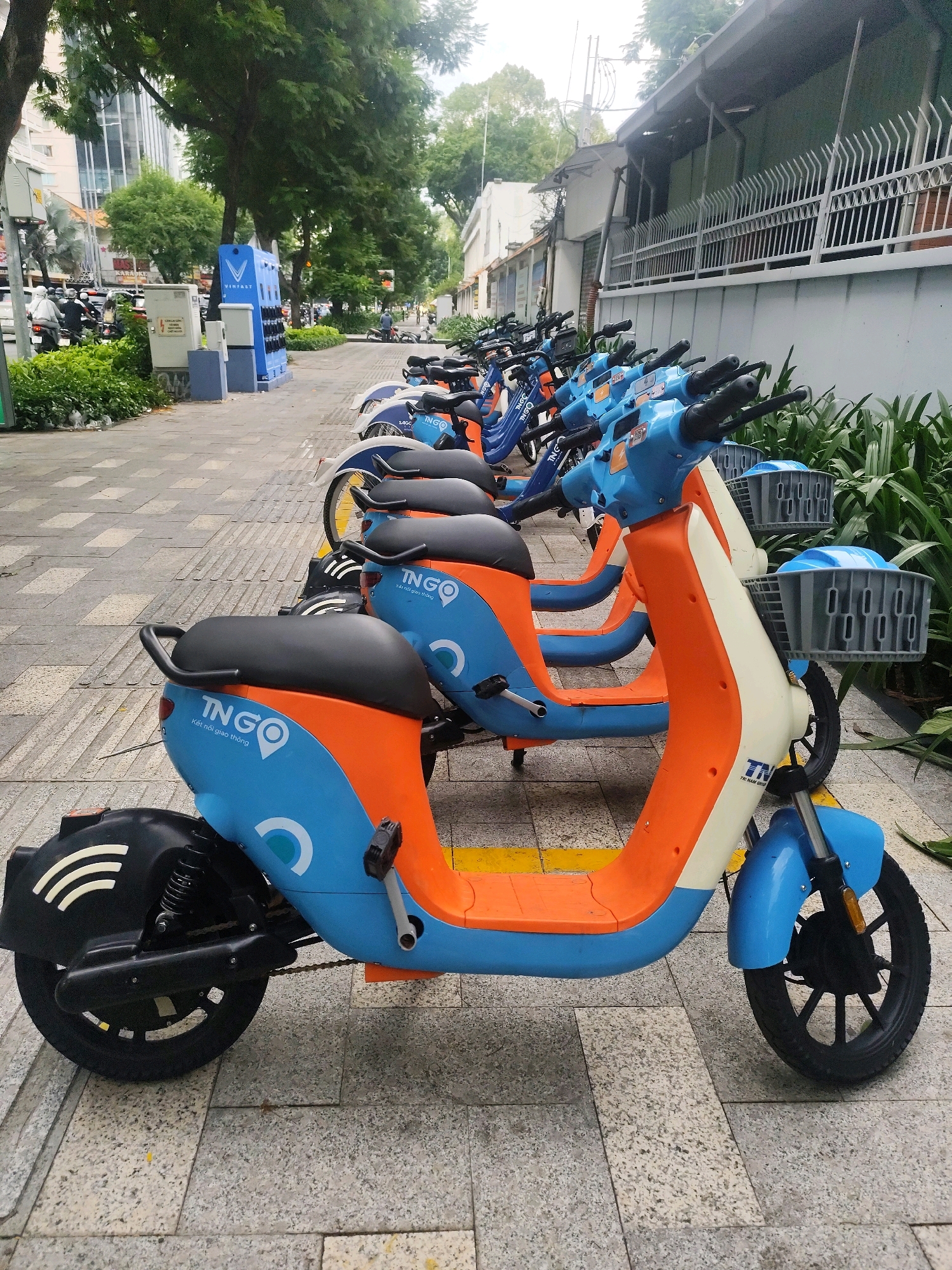 In Ho Chi Minh City, Vietnam, an outdoor scene captures a row of electric scooters parked on a sidewalk. The scooters are predominantly orange and blue, with some featuring white accents. Each scooter is equipped with a basket at the front. The sidewalk is paved with grey and beige tiles, marked by yellow lines. In the background, a street is visible with traffic, including motorcycles and cars. Trees line the sidewalk, and a building with a metal fence stands to the right. A blue charging station for the scooters is prominently displayed to the left. The sky is overcast, suggesting a cloudy day. No people are actively interacting with the scooters, indicating they are parked and awaiting use.