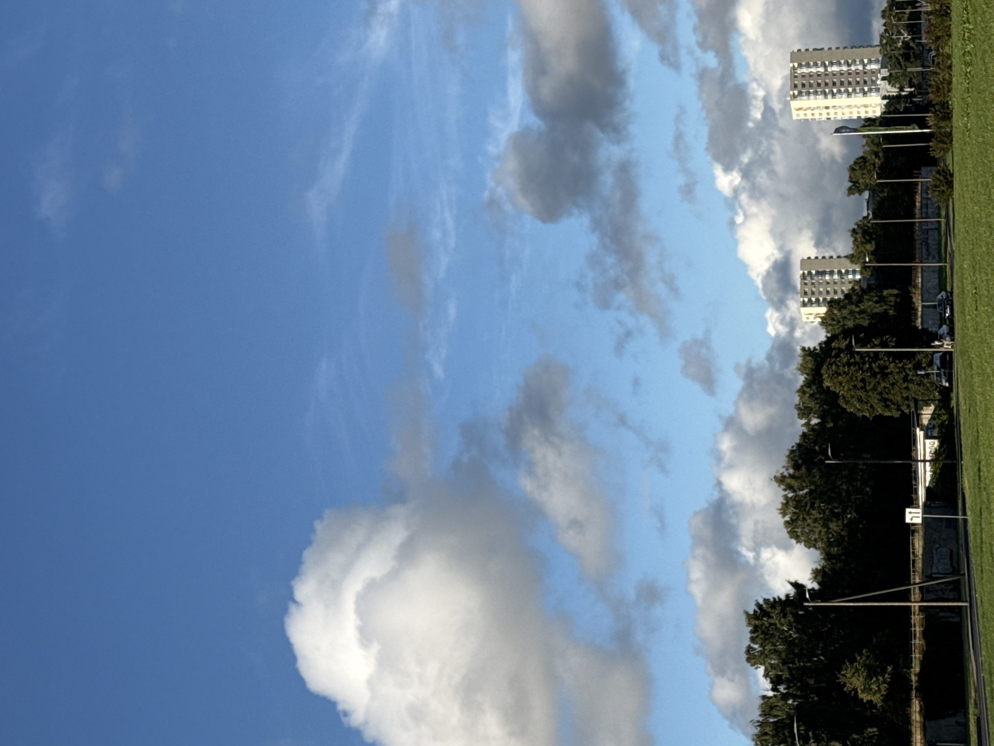 This image captures a view of the Pirita district in Tallinn, Estonia, on a partly cloudy day. The sky is a vibrant blue, with large, fluffy white clouds dominating the upper portion, interspersed with thinner, wispy clouds. In the middle ground, a dense line of dark green trees forms a natural barrier. Beyond the trees, two identical, multi-story apartment buildings stand tall. These buildings have light-colored facades and a grid pattern of windows. In the foreground, there is a paved road and a grassy area. Several streetlights are visible, along with a traffic sign indicating a turn. A few parked cars are visible on the side of the road, and two figures are walking near the parked cars. The overall scene suggests an urban residential area bordered by greenery.