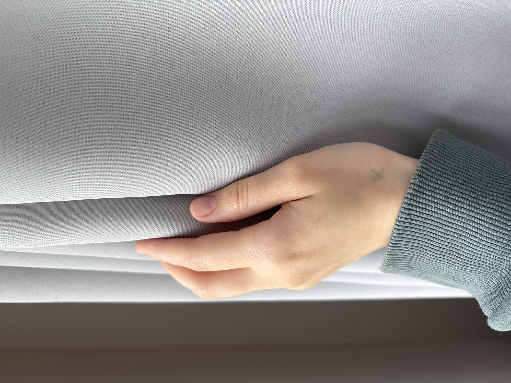 A close-up depicts a hand, identified by the light skin tone and the presence of a small, dark birthmark or mole on the wrist. The hand is pulling on a grey, folded or pleated fabric, possibly a curtain or blind. The sleeve of what appears to be a sweater or long-sleeved shirt, indicated by its ribbed texture, is also visible. The bottom portion of the image is obscured by a soft, out-of-focus area, possibly a wall or another surface. There is no location information visible in this image, and Saint Petersburg, Russia, cannot be confirmed.