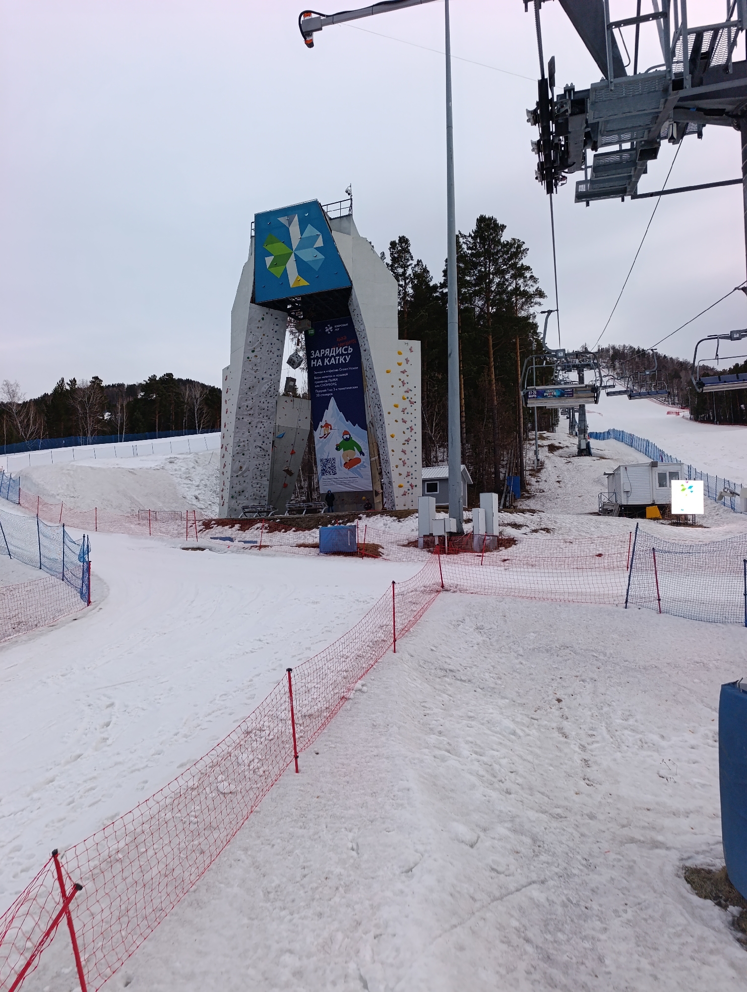 The image depicts a ski resort in Krasnoyarsk, Russia, on an overcast day. The foreground is covered in snow, with red safety netting delineating boundaries. In the middle ground, a climbing wall structure with a blue and white sign featuring a geometric design is visible. A banner advertising "Зарядись на Катку" (Charge to the Ice Rink) hangs on the climbing wall. A ski lift, with unoccupied chairs, rises uphill to the right of the climbing wall. In the background, coniferous trees line the top of a hill. A small white structure is positioned near the base of the ski lift, and signage is present to the right of it. The scene appears to be a ski area, though not actively in use at the time of the photograph.